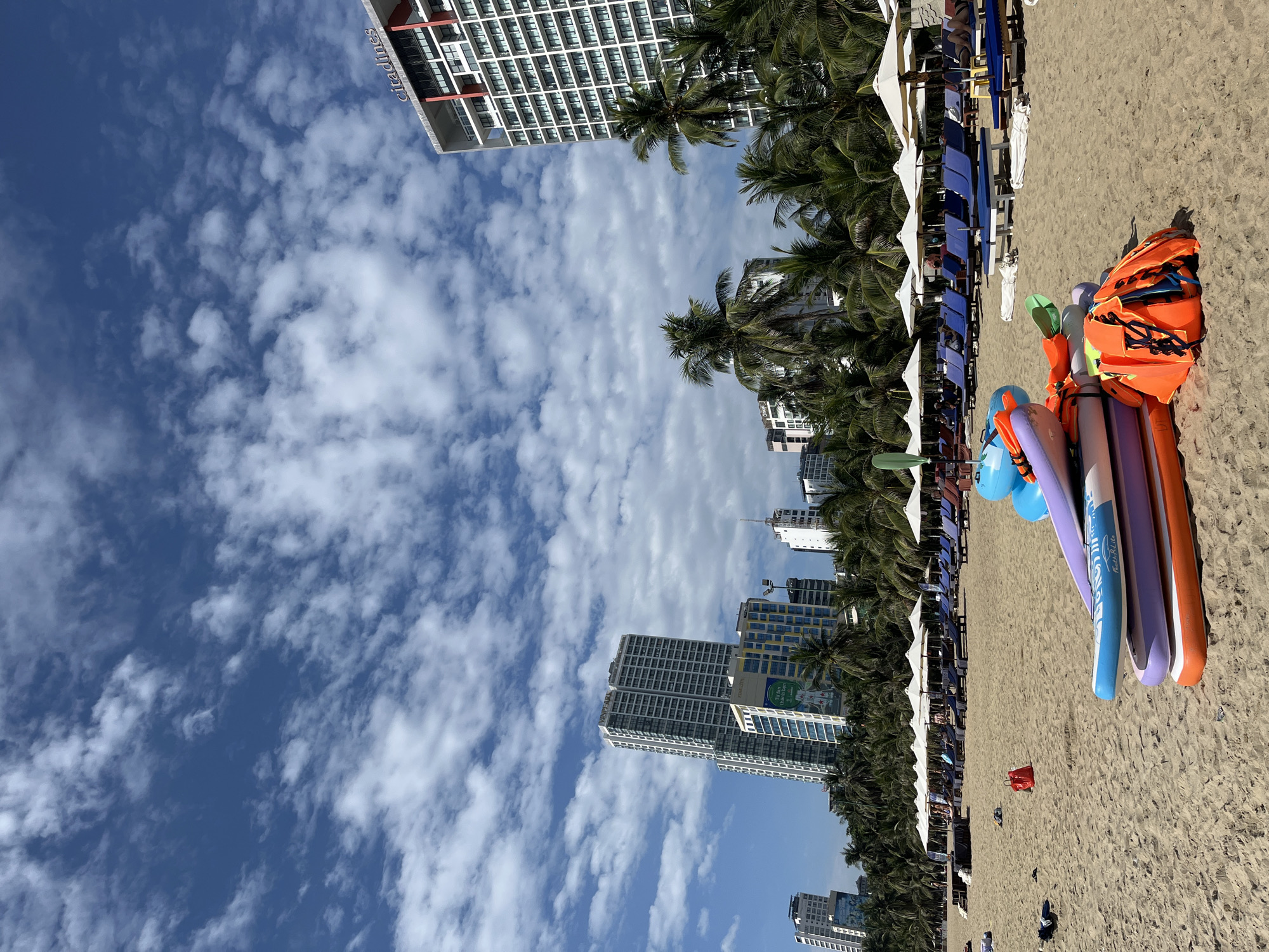 This image depicts a scene on a sandy beach in Nha Trang, Vietnam. In the foreground, several inflatable paddleboards are stacked on the sand, accompanied by orange life jackets and a green paddle. The beach is lined with rows of lounge chairs and white umbrellas, some of which are occupied by beachgoers. Palm trees and several high-rise buildings, including the Citadines hotel, are visible in the background, indicating an urban coastal setting. The sky is a vibrant blue with scattered white clouds. The overall impression is a bustling resort beach on a sunny day.