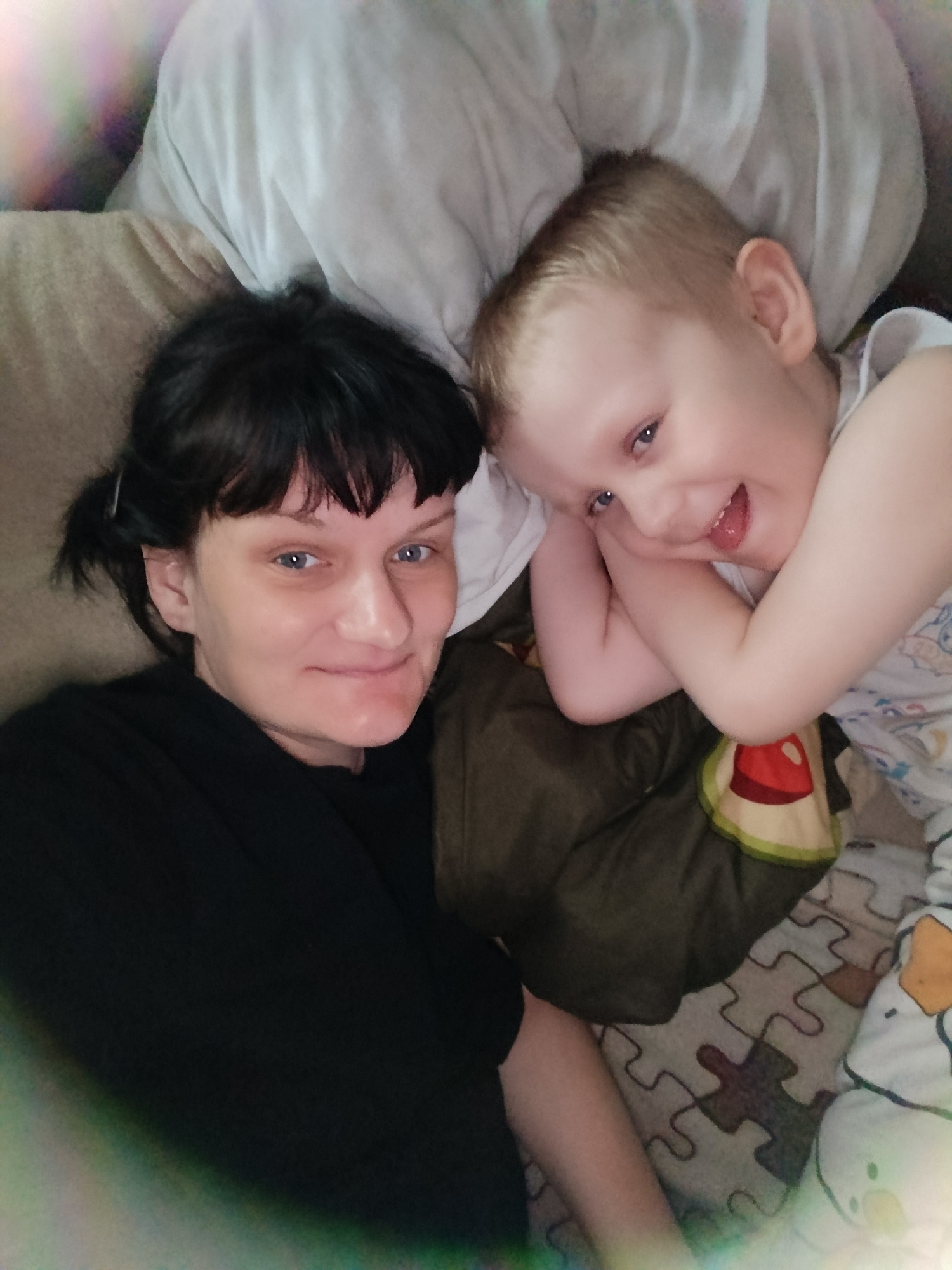 A woman and a young child are lying down. The woman has dark hair and is wearing a black shirt. The child has blonde hair and is wearing a white patterned shirt. They appear to be on a bed or couch with pillows. The bedspread has a puzzle piece pattern. The location is Unknown, Unknown.