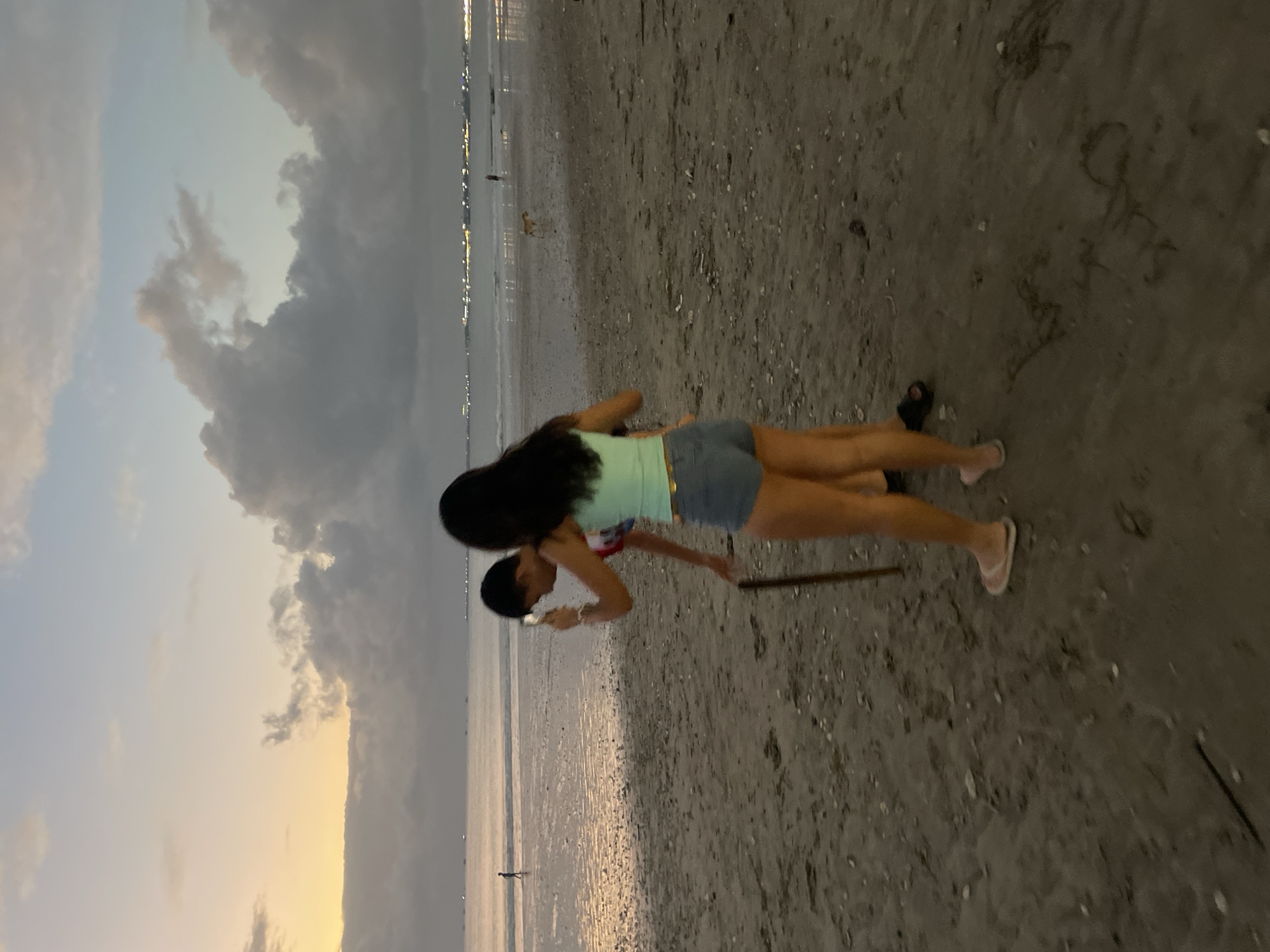 This media file shows two people on a beach at what appears to be dusk or dawn. The sky is filled with dramatic clouds, transitioning from a pale yellow and orange on the horizon to darker grays and blues higher up. The sea is calm and reflects the soft light.
In the foreground, a person is lying on their stomach on the sand, looking towards another person who is standing and leaning over them. The standing person has long, dark hair and is wearing a light blue top and denim shorts. The person lying down appears to be wearing a red and white shirt. A stick or piece of driftwood is lying on the sand between them, and it looks like the standing person might be holding it or interacting with it. The sand is damp and shows some ripples from the receding tide, with small shells and debris scattered across it.
In the background, the shoreline is visible with a line of lights in the distance, suggesting a town or city. There is a single, small figure walking in the shallow water and a dog further up the beach. The overall mood is serene and peaceful, with the soft lighting and the quiet beach setting contributing to this feeling. The time of day is indicated by the color of the sky, and the location is clearly a beach. No text is visible in the image.