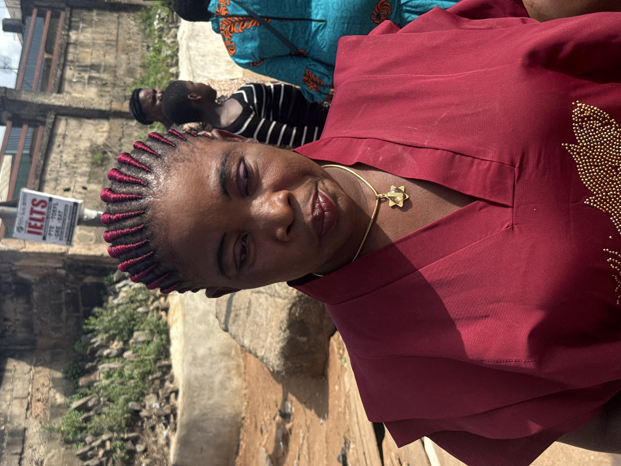 In Onitsha, Nigeria, a woman wearing a maroon-colored top with gold bead embellishments on the right side and a gold necklace stands outdoors. She has dark braided hair with red extensions at the roots. In the background, there's an IELTS signpost, indicating an educational center for English language testing. Behind her, there are other people, including one in a turquoise and orange patterned fabric and another in a black and white striped top. The environment is urban, showing buildings and some greenery. The ground appears to be a combination of dirt and possibly cracked pavement.