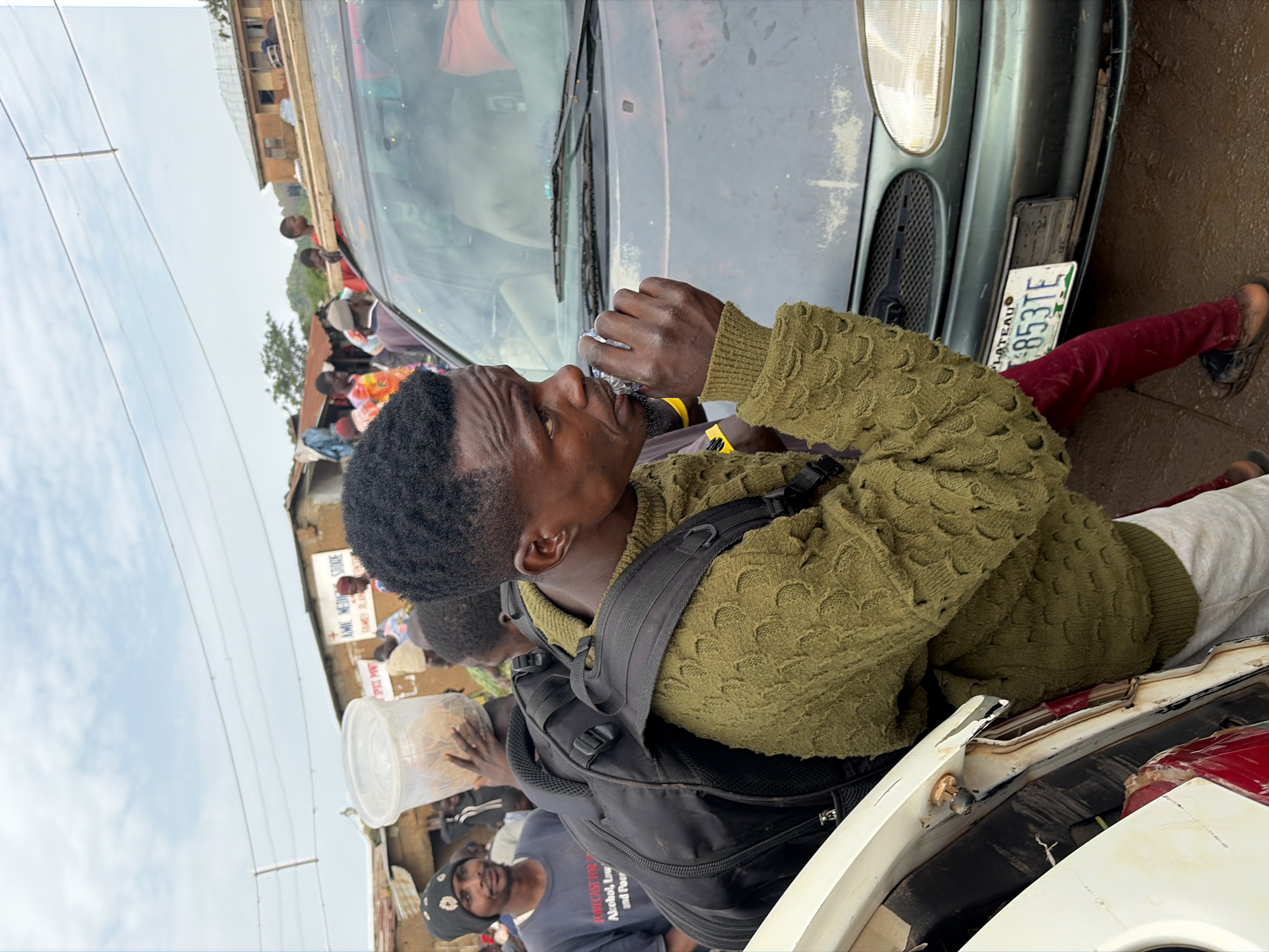 The image depicts a bustling outdoor scene, likely a street or marketplace, in Gura Top, Nigeria. In the foreground, a young Black man wearing a distinctive olive green knit sweater and a backpack is the central focus. He is seen drinking from a small, crinkled wrapper held in his right hand, his gaze directed to his right. Beside him, to his left, is the rear of a white car, partially obscuring a red object within it. To the right of the man is a dark green sedan, its front end visible, including a license plate that reads "PLATEAU L853TE." The background reveals a crowded environment with numerous people milling about. Structures that appear to be shops or homes line the street, some with corrugated metal roofs. Signs are visible on some buildings, including one with red lettering that seems to say "ISLAMIC MEDICAL STORE." Overhead, power lines crisscross the sky, indicating an urban or semi-urban setting. The ground appears to be unpaved, with some wet patches visible. The overall atmosphere suggests a lively and somewhat chaotic daily life.