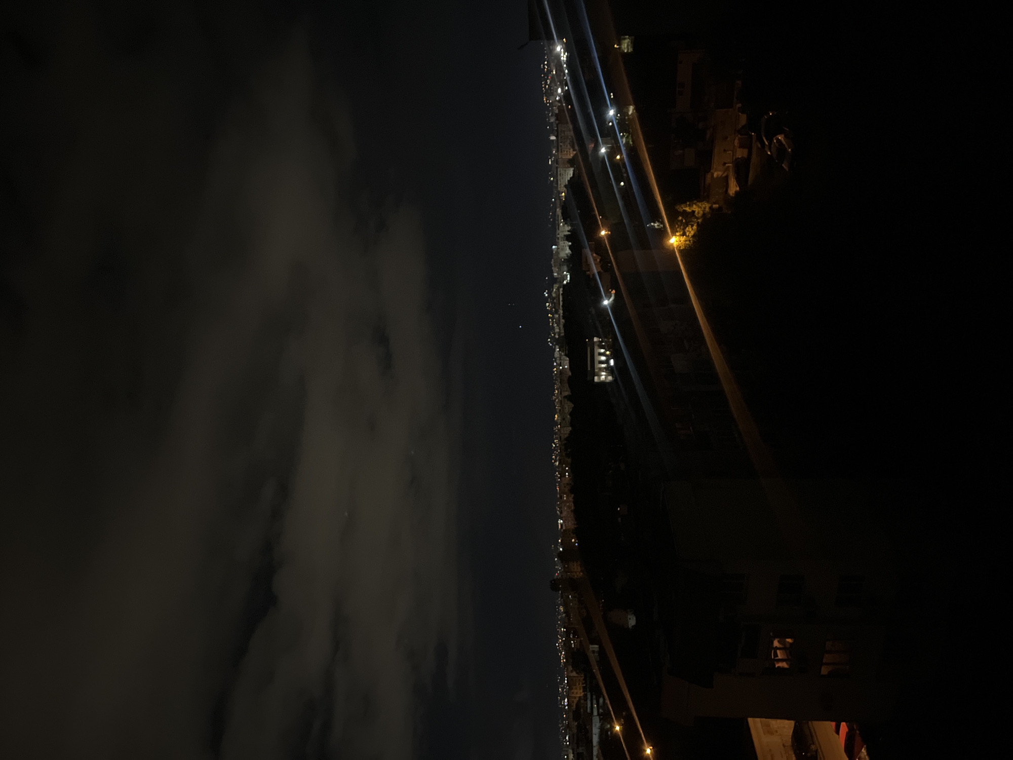 The image depicts a nighttime cityscape of Sofia, Bulgaria. On the left side, there are dark, cloud-covered skies. The right side of the image features a skyline with multiple lit buildings and streetlights. The foreground is dark, with some obscured building structures and possible street furniture visible. Overall, it's a dimly lit scene, suggesting night or late evening.
