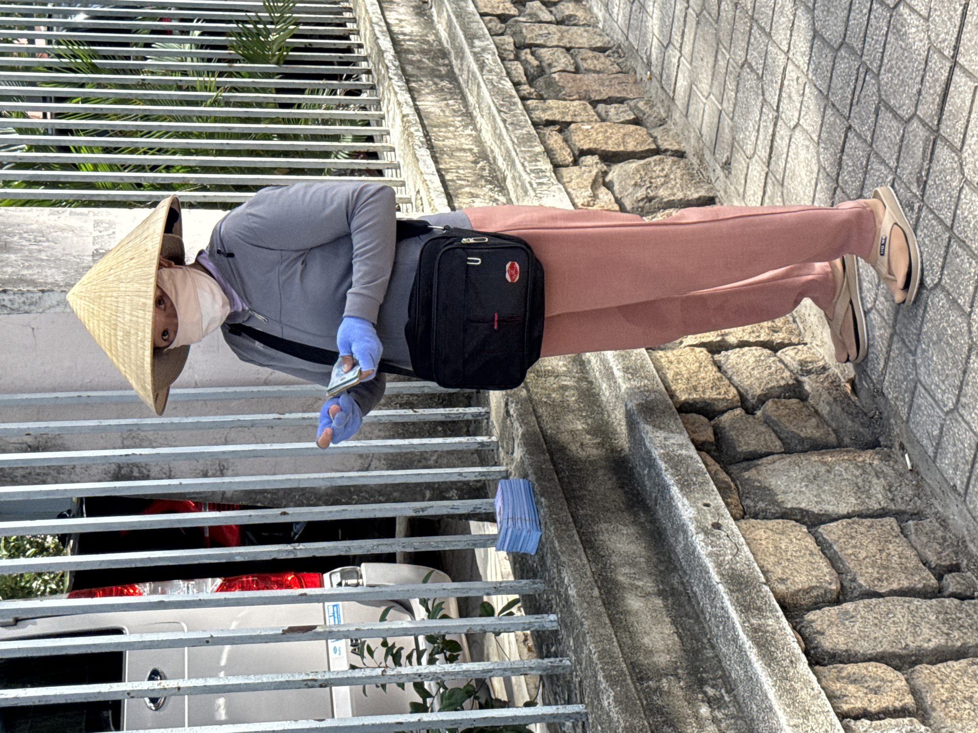 A person wearing a conical hat, a light pink face mask, a grey hooded jacket, and pink trousers stands on a sidewalk. The person is wearing blue gloves and holding a stack of bills in their hands. A black crossbody bag hangs from their shoulder. To the left of the person, a stack of blue-wrapped bills sits on a low stone wall. Behind the wall is a metal fence, and through the fence, a silver van is visible. The scene appears to be a street in Nha Trang, Vietnam, given the presence of the conical hat, the style of the van, and the overall urban environment. The ground is paved with stone tiles, and a stone wall with a concrete cap runs along the sidewalk. Sunlight casts shadows on the ground, indicating it is daytime.