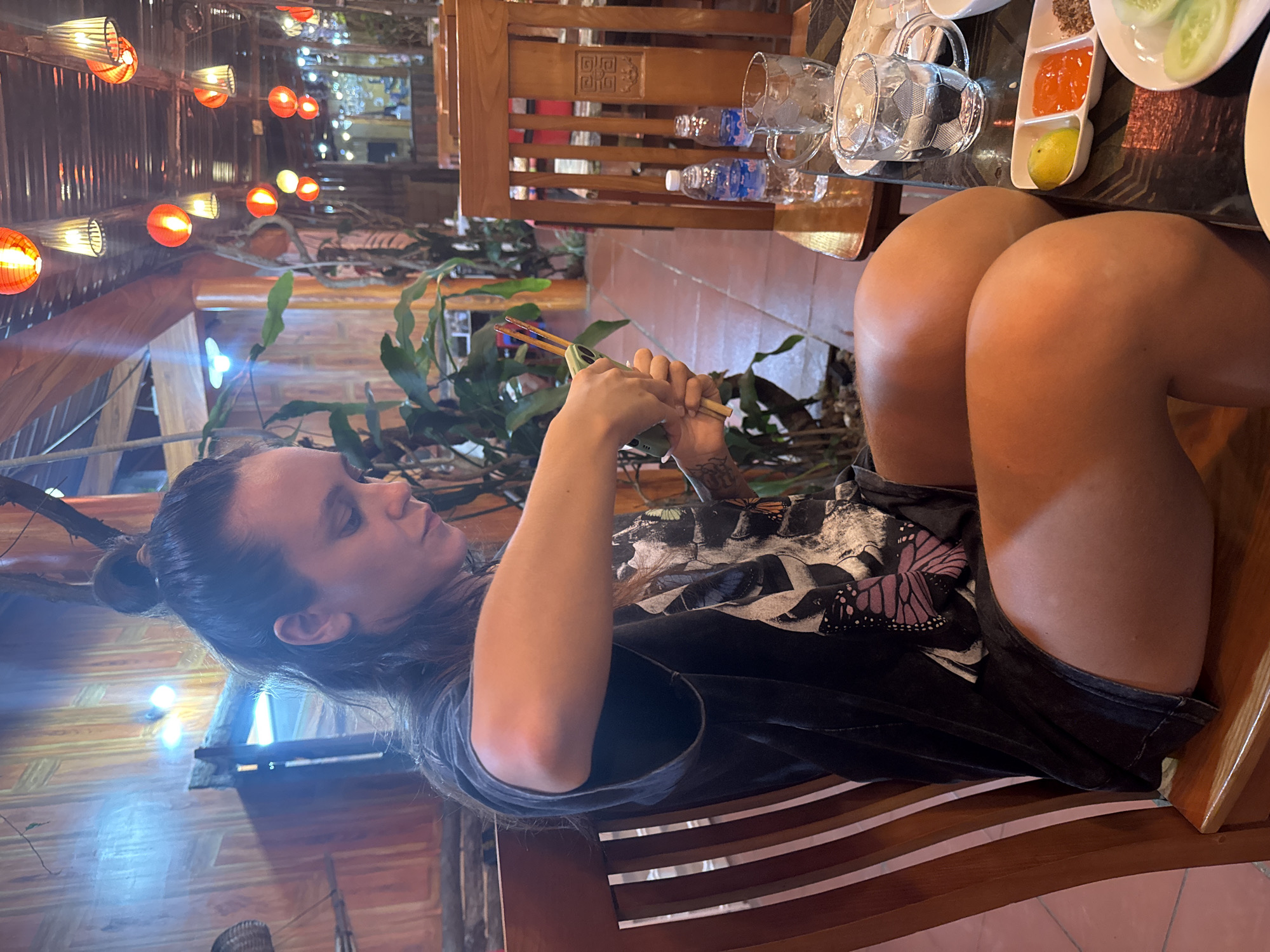 The image shows a woman seated at a wooden table in a restaurant in what appears to be the Mèo Vạc Commune, Vietnam, based on the traditional architecture and decor. She is using chopsticks. On the table are plates with food, along with water bottles and glasses. The restaurant has traditional lanterns hanging from the ceiling. The woman is wearing a dark-colored t-shirt with a butterfly design.