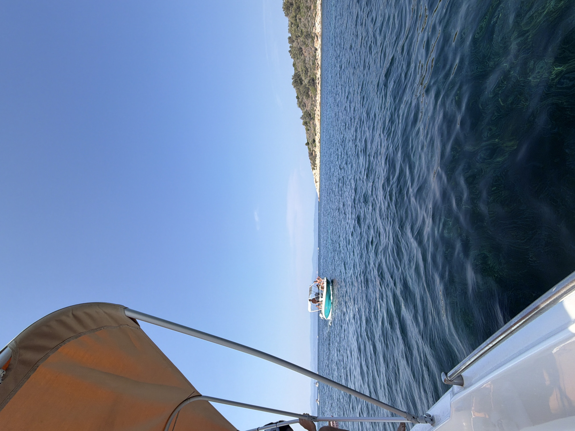 A long shot captures a view from a boat at sea under a clear blue sky. In the foreground, the tan canopy and silver railings of the boat are visible, partially obscuring the scene. The sea is a deep blue, with ripples on its surface. A small turquoise boat with a white canopy is in the middle distance, carrying several people. A forested island with rocky shores is visible on the horizon. The image is likely taken in Greece, possibly on a sunny day.