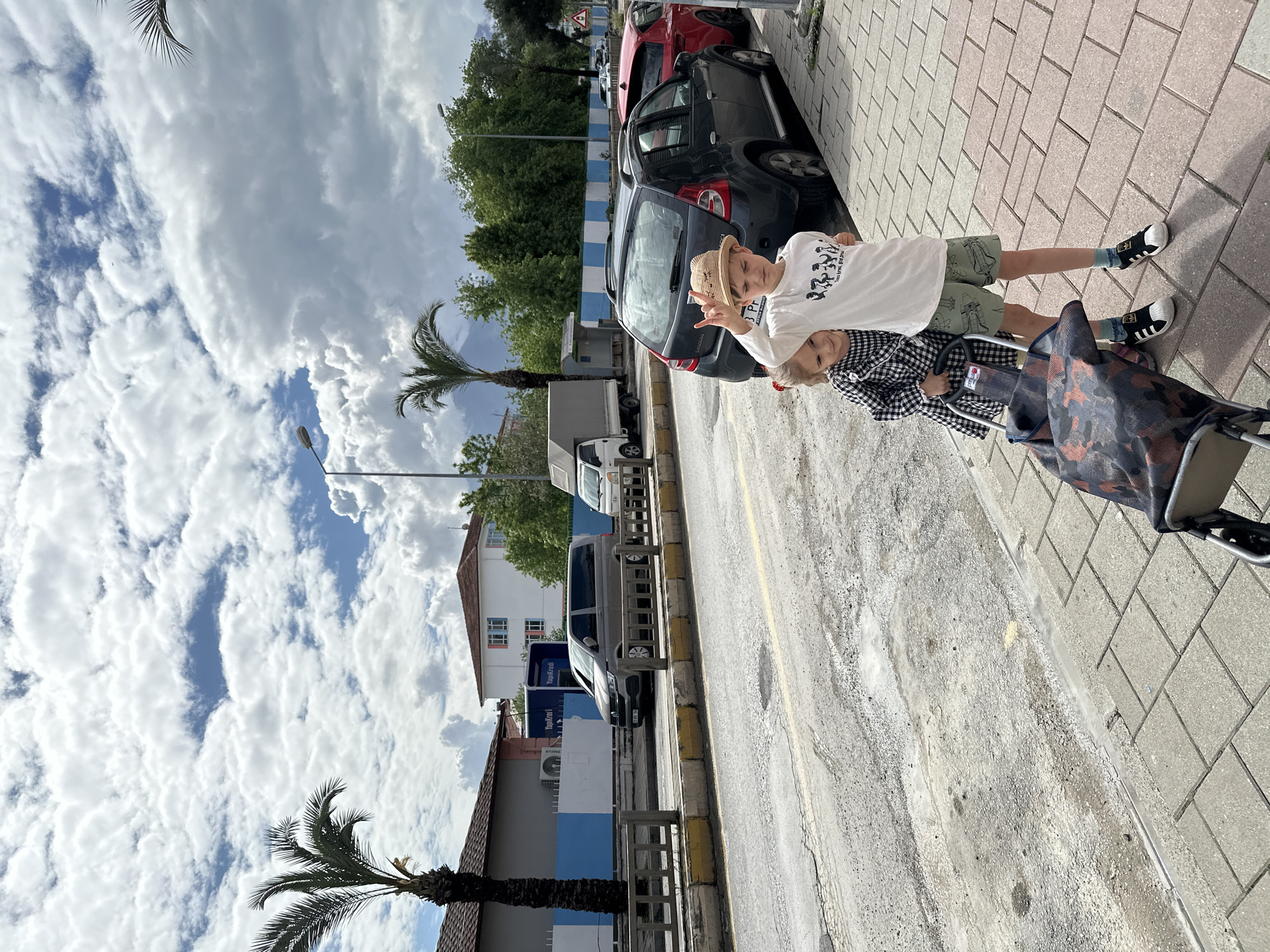 On a sunny day in Fethiye, Turkey, two children stand on a sidewalk next to a street lined with parked cars. The older child, wearing a straw hat, white t-shirt, and green shorts, makes a "peace" sign with their fingers. The younger child, dressed in a black and white checkered shirt, holds onto a shopping cart. A palm tree is visible in the foreground, while buildings and trees line the street in the background. The sky is filled with fluffy white clouds.