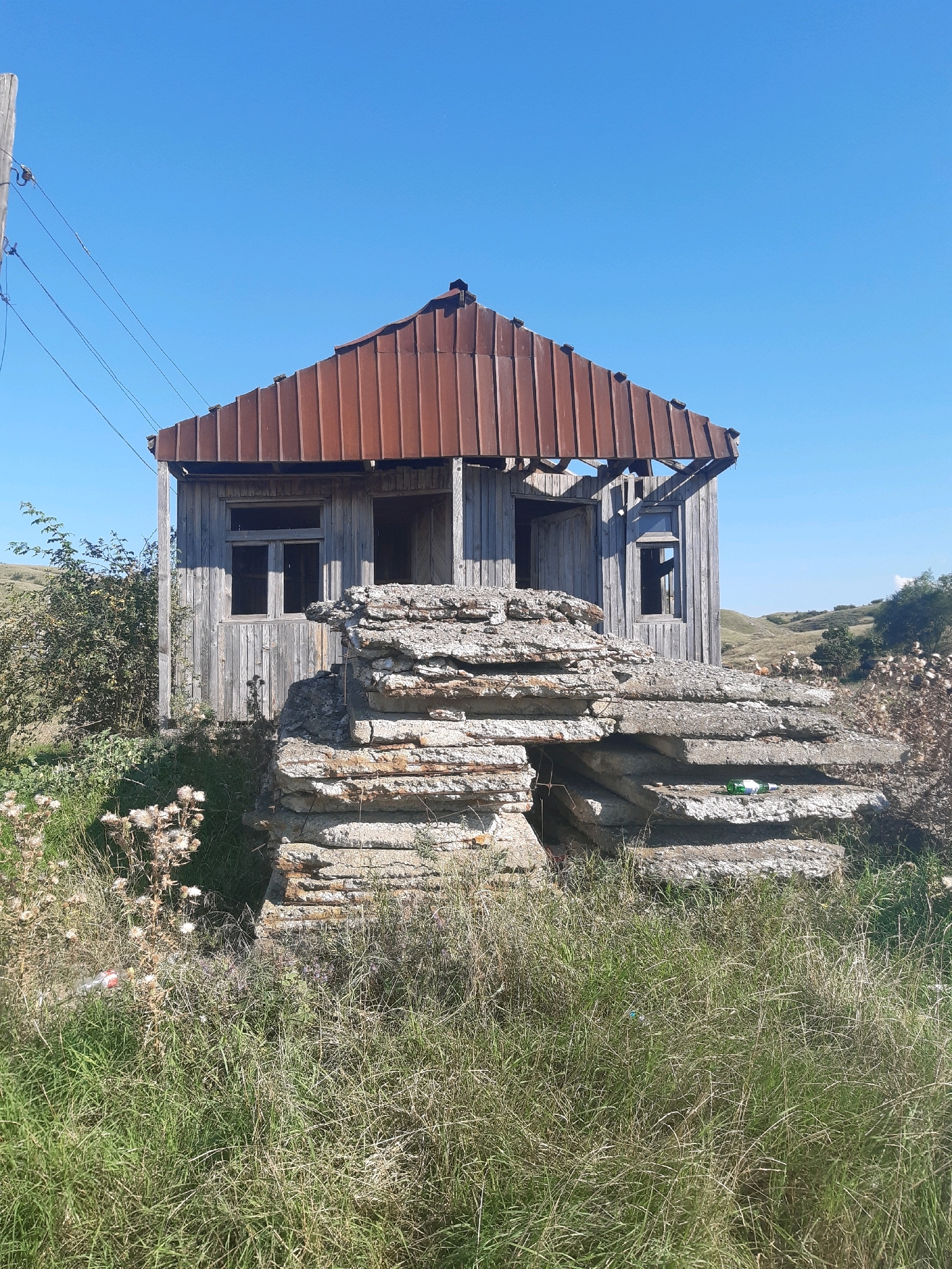 An abandoned wooden structure with a rusted metal roof stands in a field in Mepiskalaki, Georgia. The structure is weathered and partially open to the elements, with visible structural damage. In front of the building, several large, flat concrete slabs are stacked precariously, forming a rough mound. The surrounding area is covered in dry grass and weeds, with some scrub brush to the left of the building. In the distance, rolling hills are visible under a clear blue sky. A wooden utility pole with wires is positioned to the left of the frame, near the building. There are no people visible in the image.
