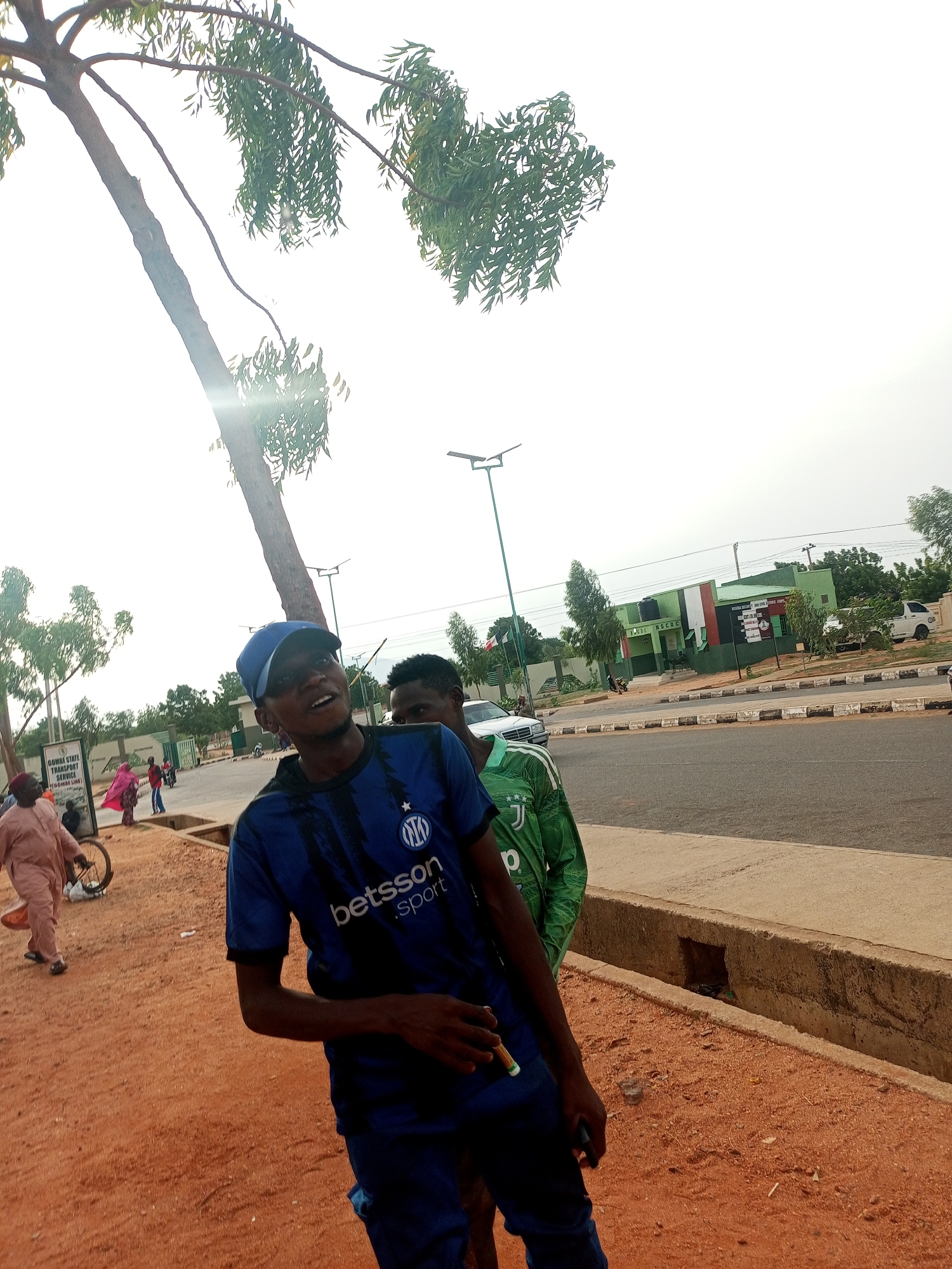 The photo captures a street scene in Gombe, Nigeria. In the foreground, two young men stand next to a concrete-lined drainage ditch. The first man wears a blue soccer jersey and a blue cap, holding a small white object. The second man stands behind him wearing a green soccer jersey. A road runs parallel to the drainage ditch. Across the road, a building with green and white exterior walls is visible. Signage on the building displays "NSCDC NSCDC." Other trees are also nearby. In the background, more people can be seen walking. The sky is overcast.