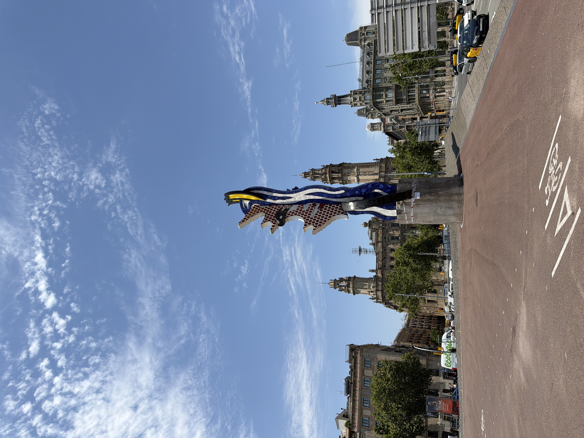 The image shows the sculpture "La Cara de Barcelona" (The Face of Barcelona) by Roy Lichtenstein, located in front of the Hotel Colón on Avinguda de la Catedral in Barcelona, Spain. The sculpture, a colorful and stylized human face, stands tall against a bright blue sky dotted with wispy white clouds. In the background, classic European architecture with ornate facades and towers lines a wide street. A red-colored paved area, possibly a pedestrian walkway or bike lane, is in the foreground, marked with white lines. Several cars, including yellow taxis, are visible on the street. Trees and buildings are scattered throughout the urban landscape, indicating a daytime scene in a bustling city. There are no visible people in the immediate vicinity of the sculpture.