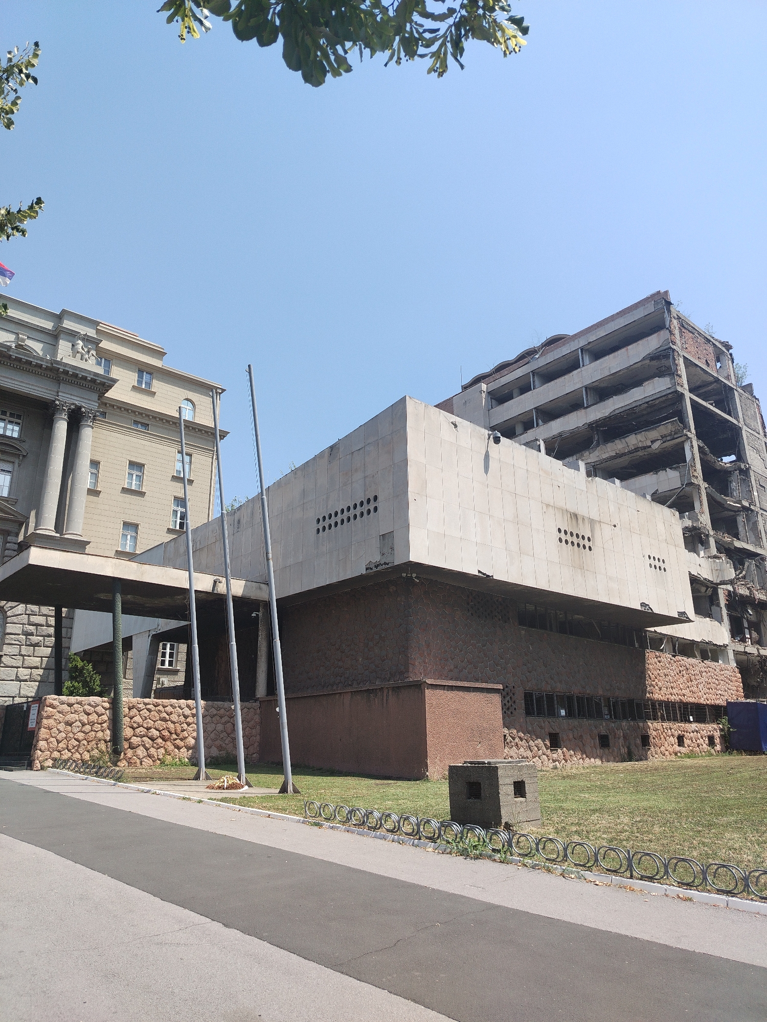 The image shows a view of the former Yugoslav Ministry of Defence building in Belgrade, Serbia, which was bombed in 1999. The building is heavily damaged, with exposed rebar and crumbling concrete. In the foreground, a paved pathway runs alongside a grassy area. Several flagpoles stand near the center of the image. To the left, a more intact building with classical columns is visible. The sky is clear and blue, and foliage from trees is present at the top of the frame.