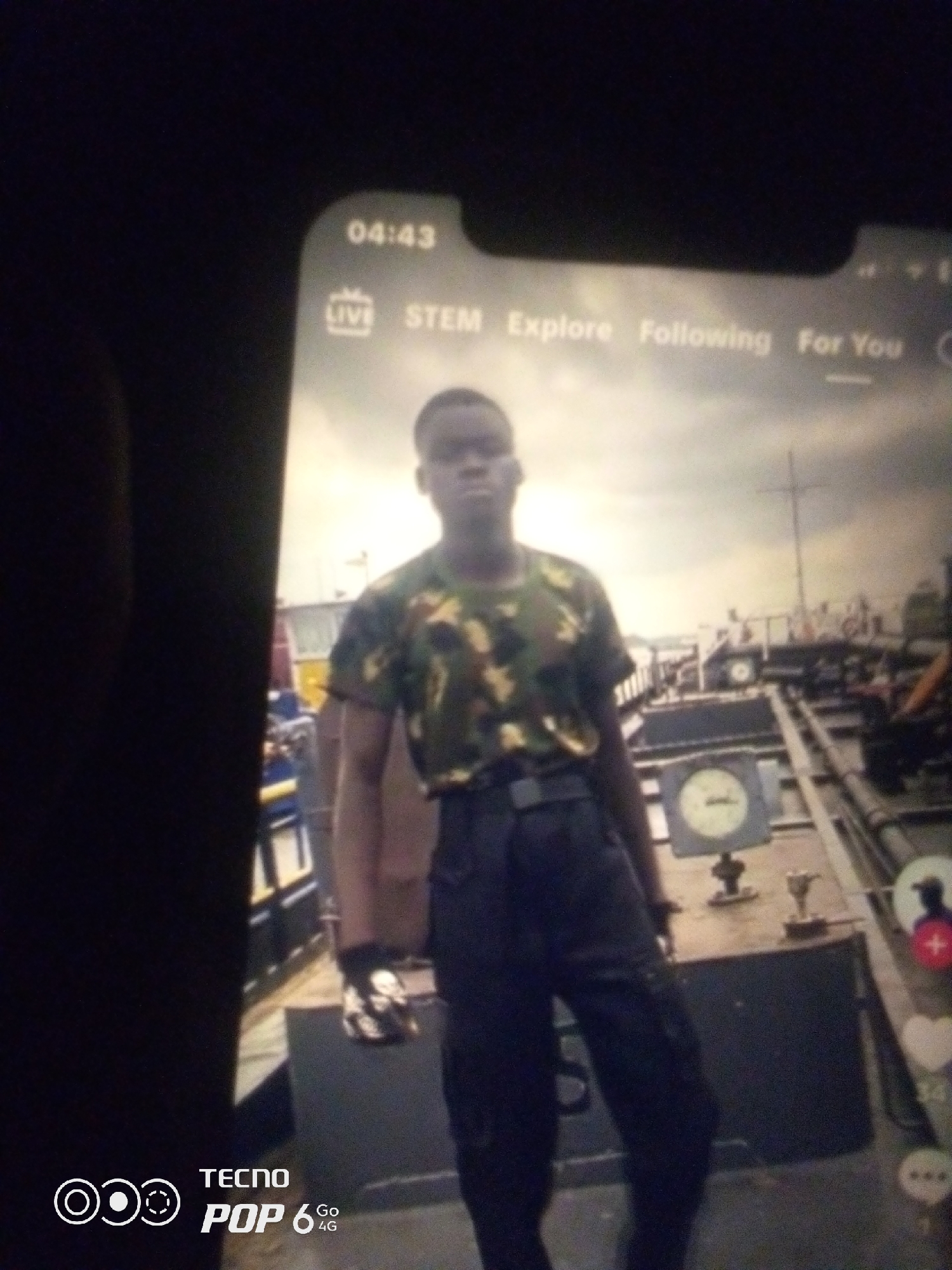 A young Black male in a camouflage shirt and dark pants stands on what appears to be a deck of a ship. He is looking slightly to his right. The background shows a cloudy sky and parts of the ship's structure, including railings, pipes, and some machinery. On the right side of the image, there are social media icons for liking, commenting, and sharing. In the bottom left corner, the "TECNO POP 6 Go 4G" branding is visible. The image quality is poor, with low resolution and blurriness, suggesting it was taken with a mobile phone camera at a distance. There is no information in the image to suggest a specific location in the city of Howayi, Nigeria.