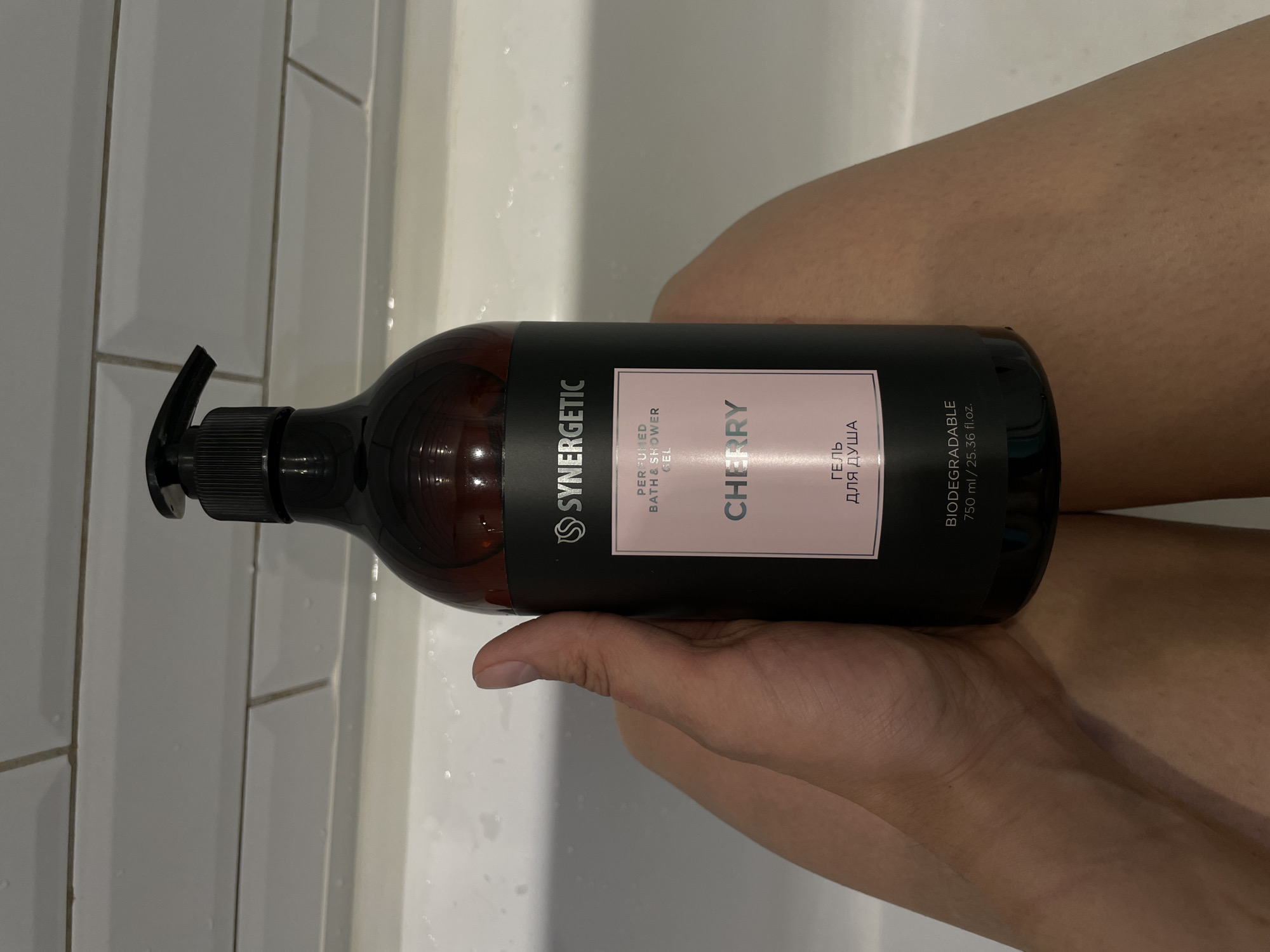 A hand holds a black bottle of Synergetic brand cherry-scented bath and shower gel. The bottle has a pink label with text in both English and Russian. The surrounding environment is a bathroom setting, with white tiles visible in the background and a glimpse of a person's leg. The image is well-lit, emphasizing the details of the product and its packaging.
