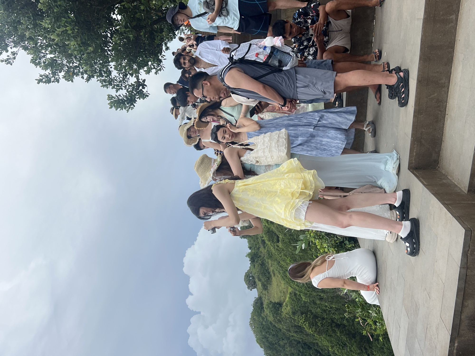 A crowded outdoor scene captures a diverse group of people, primarily tourists, gathered on a light-colored concrete or stone pathway. The image is taken from a slightly elevated perspective, looking down at the individuals and out towards a lush, green landscape under an overcast sky.
The subjects are dressed in casual, summery attire suitable for warm weather, including shorts, t-shirts, light dresses, and sun hats. Many are wearing sandals or comfortable shoes. Backpacks are visible on several individuals, and some are holding cell phones or cameras, suggesting a sightseeing activity. One woman in a yellow dress is actively using a small camera, while another woman in a white dress kneels on the ground, possibly posing for a photograph. The people appear to be waiting in a loose line or observing the surroundings.
The setting features dense, tropical green vegetation covering rolling hills in the background, characteristic of a scenic natural environment. The sky is largely overcast with soft, diffuse clouds, indicating daytime. The scene appears to be a popular viewpoint or attraction in Nusa Penida, Indonesia, given the large gathering of people and the distinct tropical landscape. No visible text is discernible on clothing or signs. The overall mood is relaxed and observant, typical of tourists exploring a beautiful location.