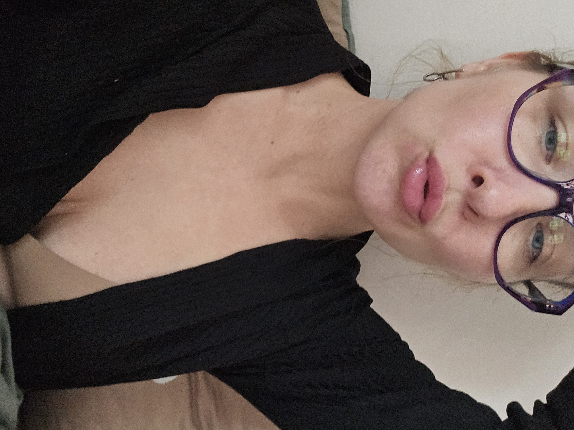 A woman is pictured close-up, wearing glasses and a black top. The location is not discernible from the image, so it's impossible to determine if it's in Sochi, Russia or any other city. The image focuses on the woman's face and upper torso. She is looking towards the camera with her lips pursed. Her skin is fair, and her eyes are blue. The top she is wearing has a ribbed texture. The background is a blurry combination of beige and white, likely indicating a room interior.