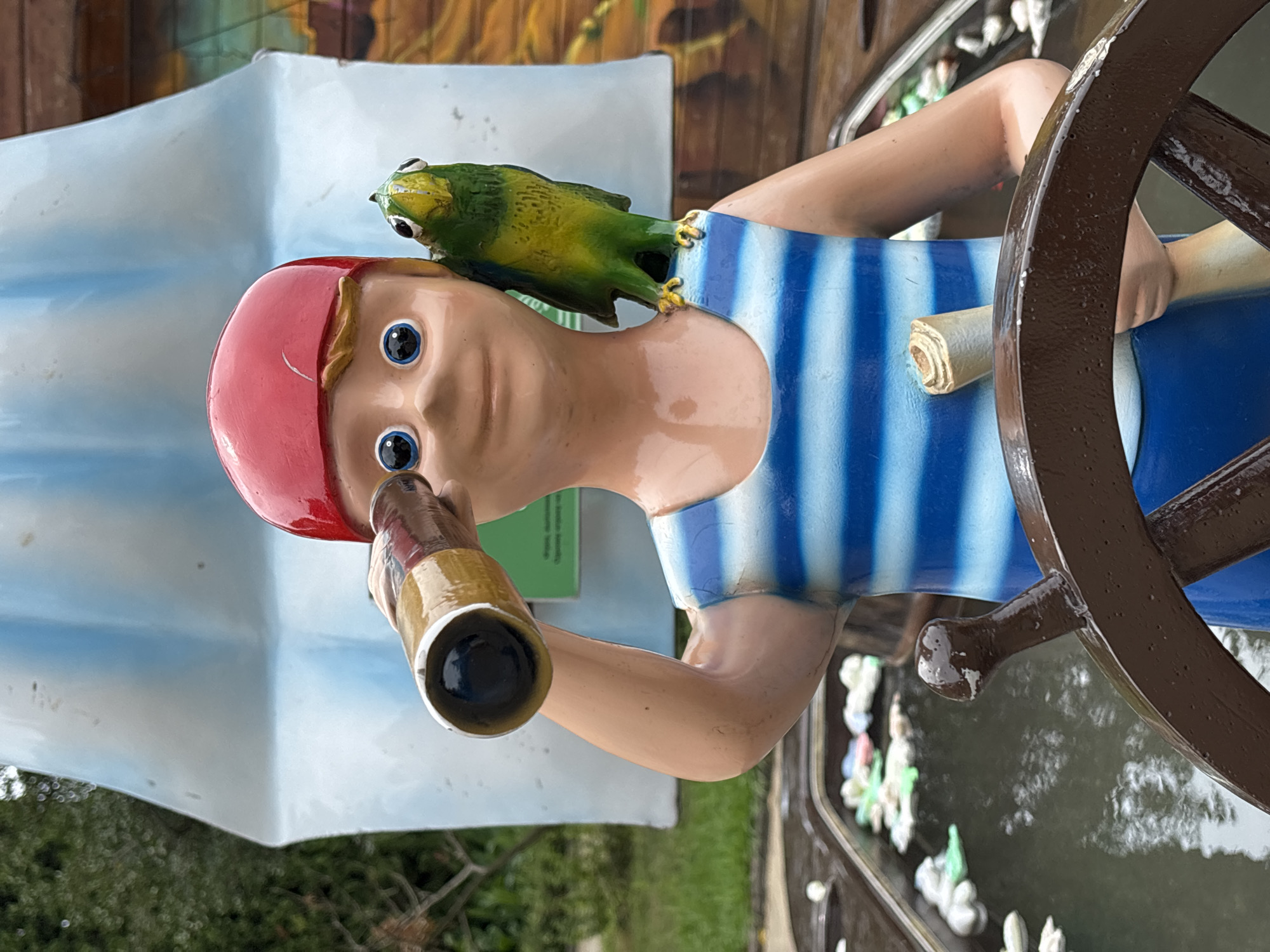 A life-sized, colorful statue of a young boy dressed as a pirate stands at a ship's helm in a water feature. The boy wears a red pirate hat, a blue and white striped shirt, and holds a spyglass to his right eye with his left hand. A green parrot is perched on his left shoulder. The statue is positioned in front of a backdrop depicting a ship's sail and a colorful mural. The water feature contains several small, decorative white and green objects floating on the surface, resembling lily pads or small boats. The scene is outdoors with greenery visible in the background. The statue and its setting appear to be part of a recreational area or attraction, possibly a park or amusement area. The location is within the city of Sochi, Russia.
