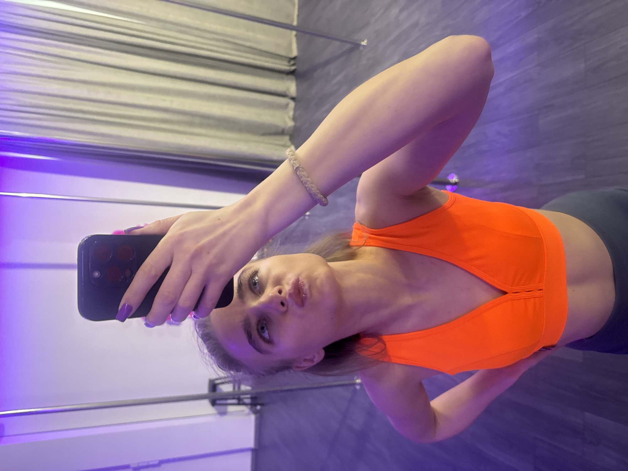 A young woman is taking a selfie indoors in a location that appears to be a dance studio or gym. She is wearing an orange sports bra and dark-colored shorts or leggings. Her right arm is extended, holding a black smartphone with a visible triple-lens camera, positioned to capture the shot of herself. Her left arm is bent and partially visible in the lower left corner. She has a light skin tone, blonde hair, and blue eyes, and is puckering her lips slightly for the photo. Her fingernails are painted purple. Her left wrist has a braided bracelet. The background includes a silver metal pole, a gray wall with horizontal linear patterns, and a purple and gray floor. The lighting in the room gives off a purplish hue.