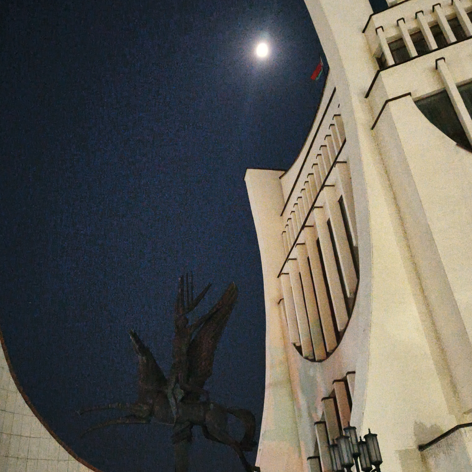 This image captures a nighttime scene in Hrodna, Belarus, at Ozheshko Street 3, depicting the Hrodna Regional Executive Committee building and the Monument to the Liberator Soldier. The upper two-thirds of the frame consist of a dark, grainy night sky, with a prominent, bright full moon positioned in the upper center. A red and green flag, consistent with the flag of Belarus, is visible on a pole projecting from the upper right section of the building.
The building itself is a multi-story structure with a light-colored (off-white/beige) facade, characterized by Soviet Modernist architectural elements including a curved main body, strong vertical fins, and dark, recessed windows. In the left foreground, a dark equestrian statue, likely bronze, portrays a figure on horseback with a raised, multi-pronged, trident-like weapon. Artificial light illuminates the building, casting some light onto the statue. A multi-lamp street light fixture is partially visible at the bottom right, positioned against the building. The lower left of the frame shows a dark, textured ground surface.