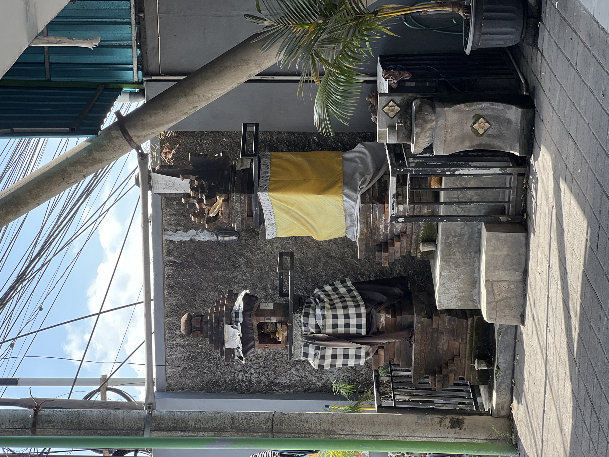 The image shows an outdoor scene in Denpasar, Indonesia, with a traditional Balinese shrine prominently featured. The shrine is adorned with two distinct cloths: a vibrant yellow cloth wrapped around one section and a black and white checkered cloth covering another. The shrine appears to be constructed from stone and brick, with multiple levels and decorative elements. A potted palm tree with lush green fronds is visible to the right of the shrine, adding to the tropical atmosphere.
In the background, a grey wall and a blue corrugated metal roof are visible. Numerous electrical wires crisscross the sky, suggesting an urban or semi-urban environment. The paving stones on the right side of the image indicate a walkway or patio. The lighting suggests it is daytime, with shadows cast by the shrine and the palm tree, indicating the sun is present. The scene appears calm and serene, with no people visible. The overall impression is of a culturally significant place, possibly a temple or a private dwelling with a shrine.