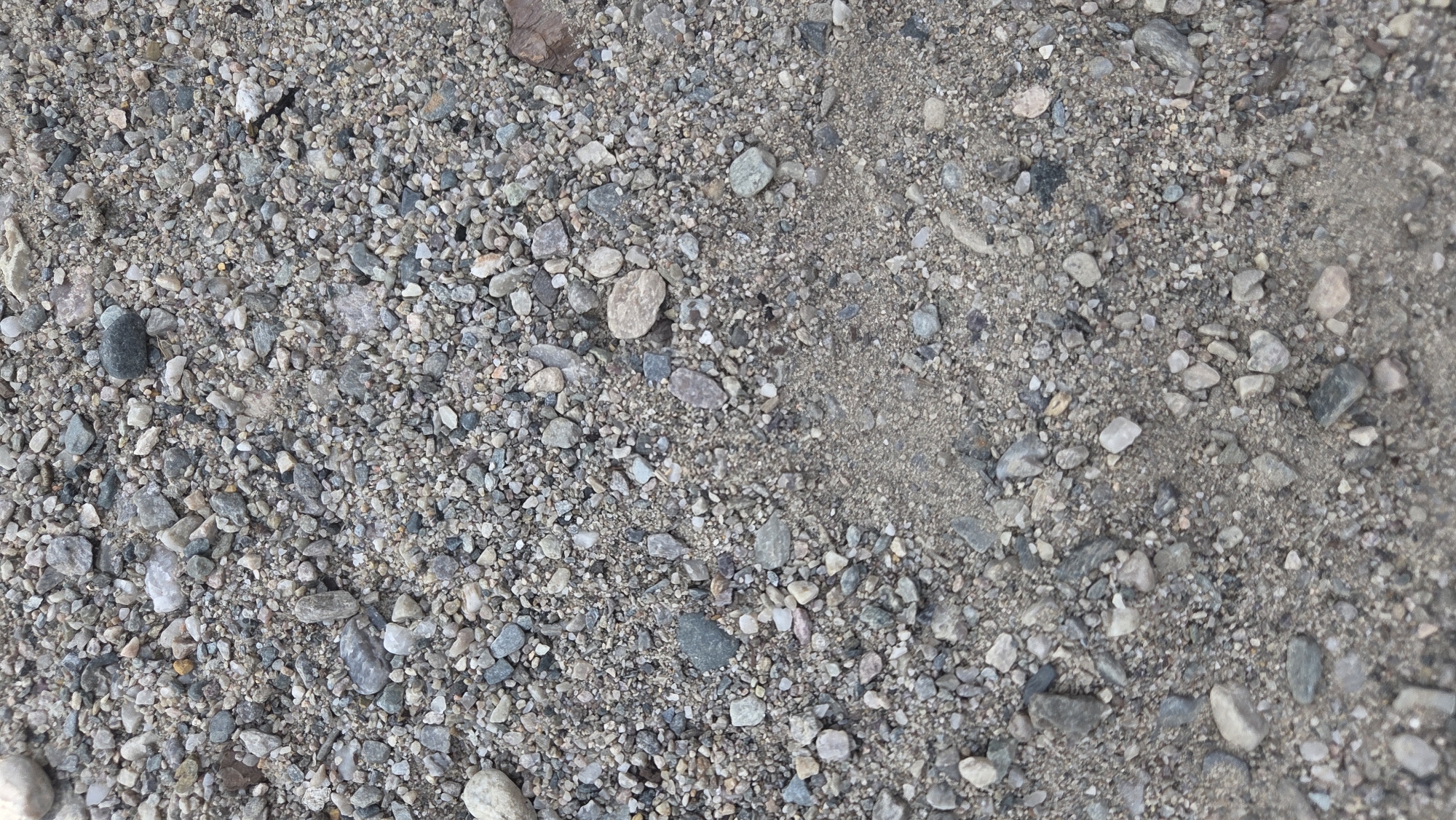 The image displays a close-up, overhead view of a ground surface composed of gravel and sand. The gravel consists of a variety of small, irregularly shaped stones of different colors, including gray, white, and brown, with some exhibiting a weathered appearance. The sand is a lighter, tan color and appears to be mixed with the gravel, with some areas showing a higher concentration of sand. The overall texture is rough and uneven. The scene is devoid of any human presence, structures, or vegetation, suggesting an outdoor, uncultivated area. Based on the prompt's instruction to include the location in Tynset, Norway, this scene likely depicts a pathway, road, or unpaved area within the city or its surrounding natural landscape.