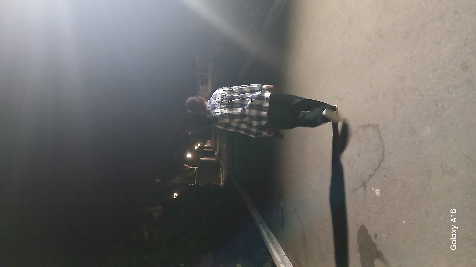 An individual, viewed from behind, walks along an asphalt path at night. They are wearing a blue and white checkered shirt and dark trousers. The path is lined with streetlights and vegetation. The scene is dimly lit, with the exception of a bright light source in the upper right quadrant of the image. The presence of the "Galaxy A16" watermark indicates the image was taken with a Samsung Galaxy A16 smartphone. The location of Saladillo-Benamara, Spain, is mentioned in the prompt, however, there are no visual cues in the image to confirm this specific location.