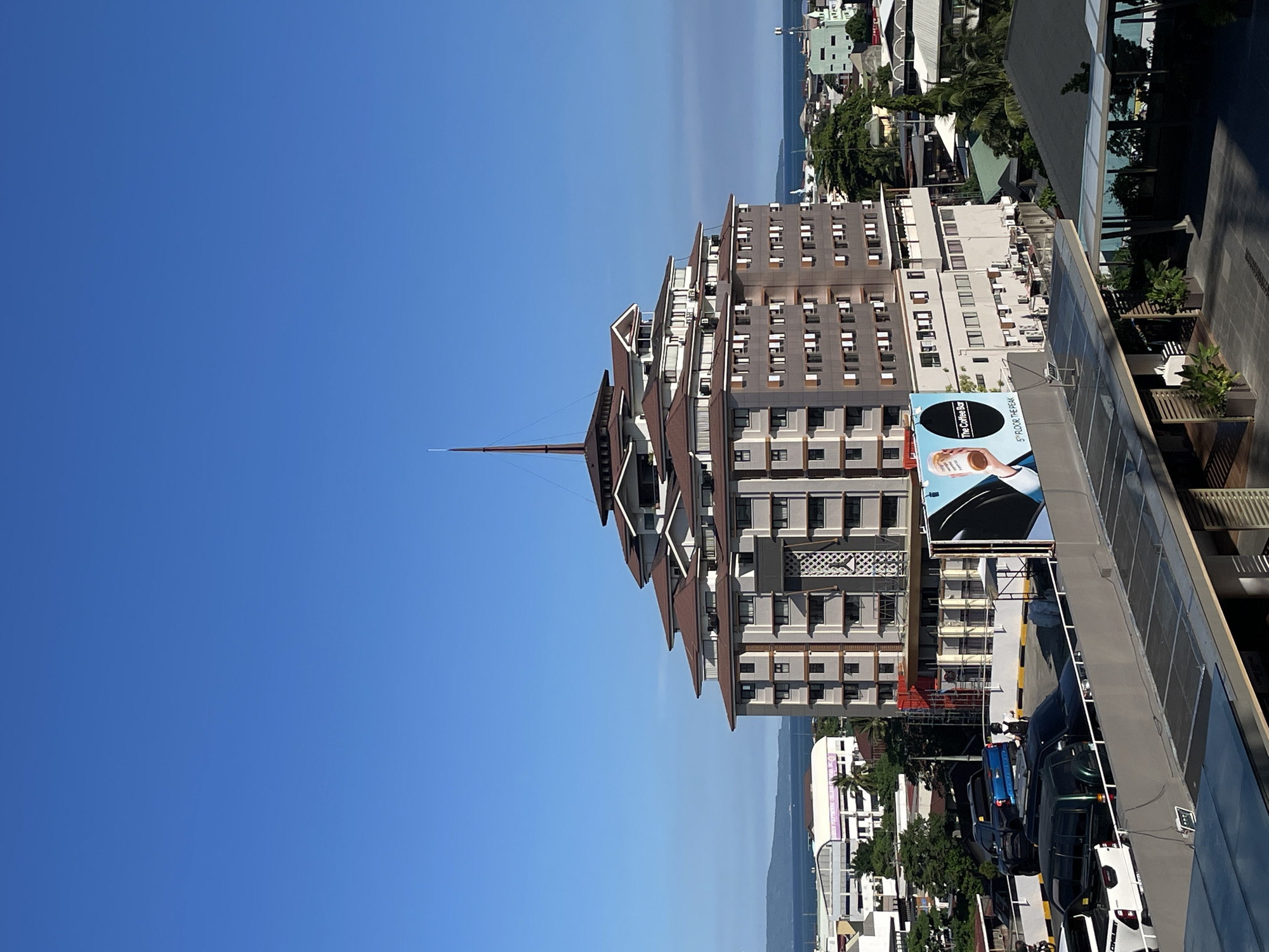 This is a tilted, high-angle shot of a city scene in Davao City, Philippines, under a clear blue sky, suggesting daytime with good weather. The primary subject is a modern, multi-story building with a distinctive tiered, pyramid-like roofline. The building's facade is composed of many windows and balconies, with a prominent antenna or spire at its peak. Adjacent to this building, to the right, is another modern structure with a glass facade, and below that, a parking lot filled with several cars.
In the background, a body of water, likely the sea, is visible, with distant hills or mountains beyond it. Other buildings, some appearing to be hotels or commercial establishments, are scattered throughout the cityscape, interspersed with greenery and palm trees, characteristic of a tropical location. One notable detail is a billboard on the side of the main building, advertising "The Coffee Bar" on the "5th FLOOR THE PEAK".
The overall impression is one of urban development and a bright, sunny day in a coastal city. There are no visible people clearly engaged in activity, and the scene is largely focused on architecture and the urban landscape.