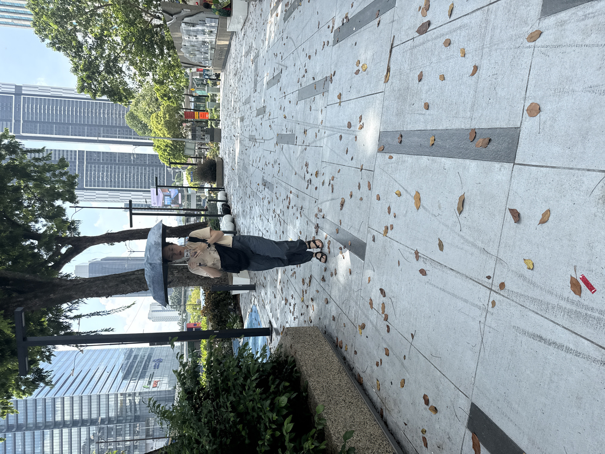 The image depicts a woman standing on a concrete tiled sidewalk in Kuala Lumpur, Malaysia, with a dark umbrella shielding her from the sun. Fallen leaves are scattered across the walkway. The woman is dressed in a light-colored shirt, dark overalls, and sandals. Skyscrapers are visible in the background, along with trees and greenery. The sidewalk's tiles alternate between light gray and darker gray stripes.