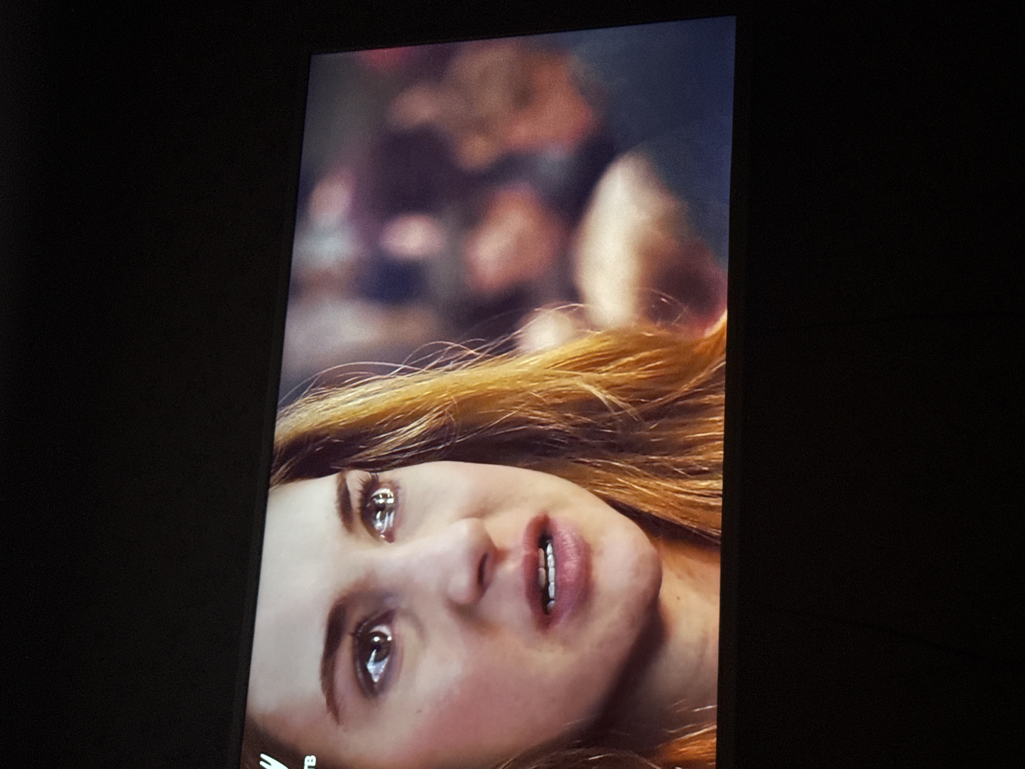 A close-up photograph shows a woman with reddish-blonde hair lying on her back. Her head is tilted slightly to the right. She has light skin, and her facial expression appears distressed. Her eyes are open, and her mouth is slightly open. The background is blurry and indistinct, suggesting other people or objects are out of focus behind her. The photograph appears to be a close-up shot taken from a screen or monitor, with a black background surrounding the image. There are no visible elements that suggest a location in Mytishchi, Russia.