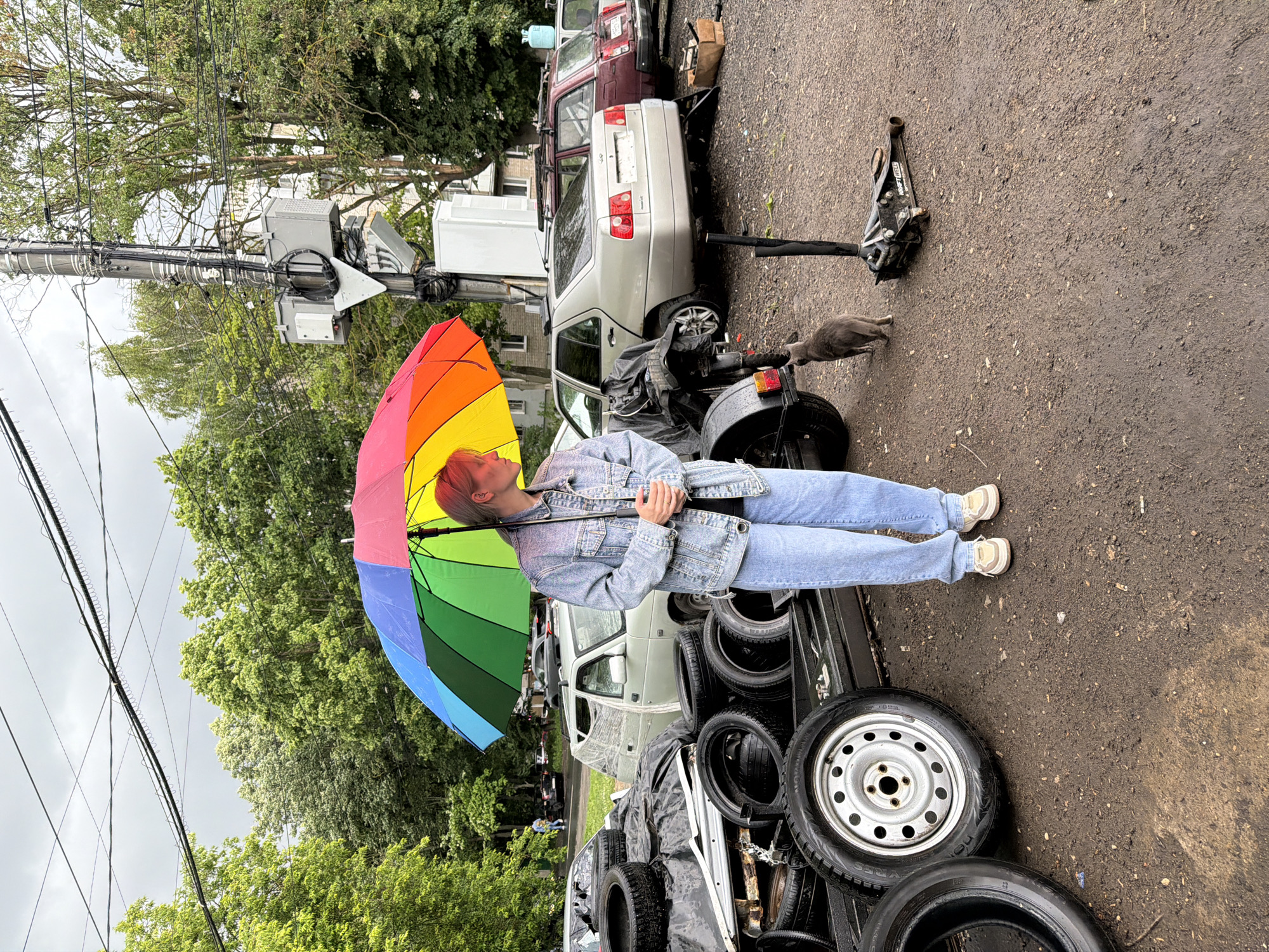 A young person stands under a rainbow-colored umbrella, holding the handle with both hands. They are wearing a denim jacket and light blue jeans. To their left is a pile of discarded car tires. Behind them, several cars are parked. A grey cat walks away from the person towards a car jack on the ground. The scene appears to be an outdoor junkyard or a car repair lot in Nizhny Novgorod, Russia, with overcast skies and wet ground suggesting recent rain. Electrical wires and a utility pole are visible in the background.