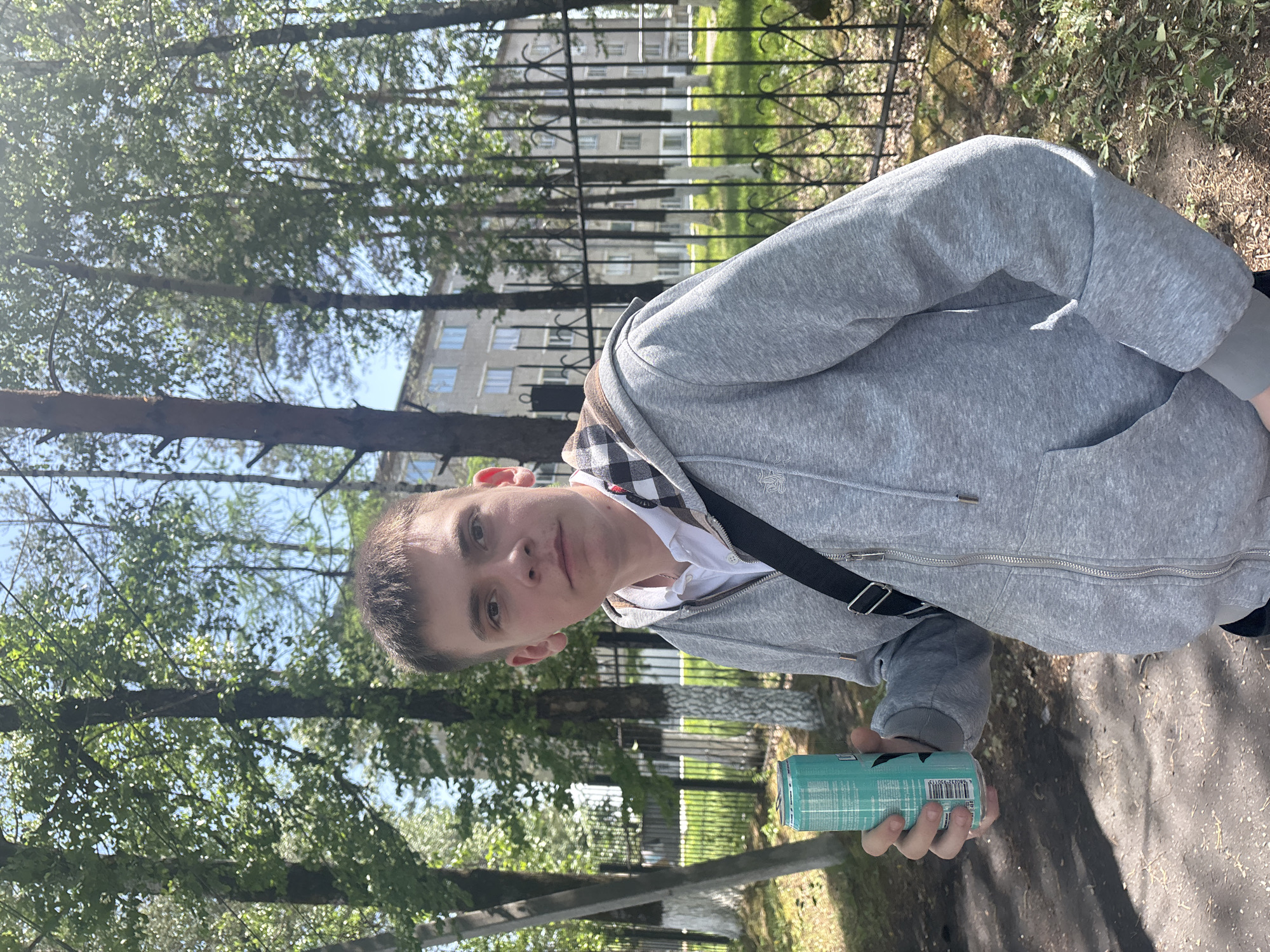 A young man is standing outside on a sunny day. He is holding a teal-colored can in his right hand. He is wearing a grey hooded sweatshirt with a black and white checkered hood, a white shirt, and a black strap across his chest. He is looking directly at the camera. The background includes trees, a metal fence, a grassy area, a paved path, and a tall building in the distance. The location appears to be a park or a green area in an urban setting.