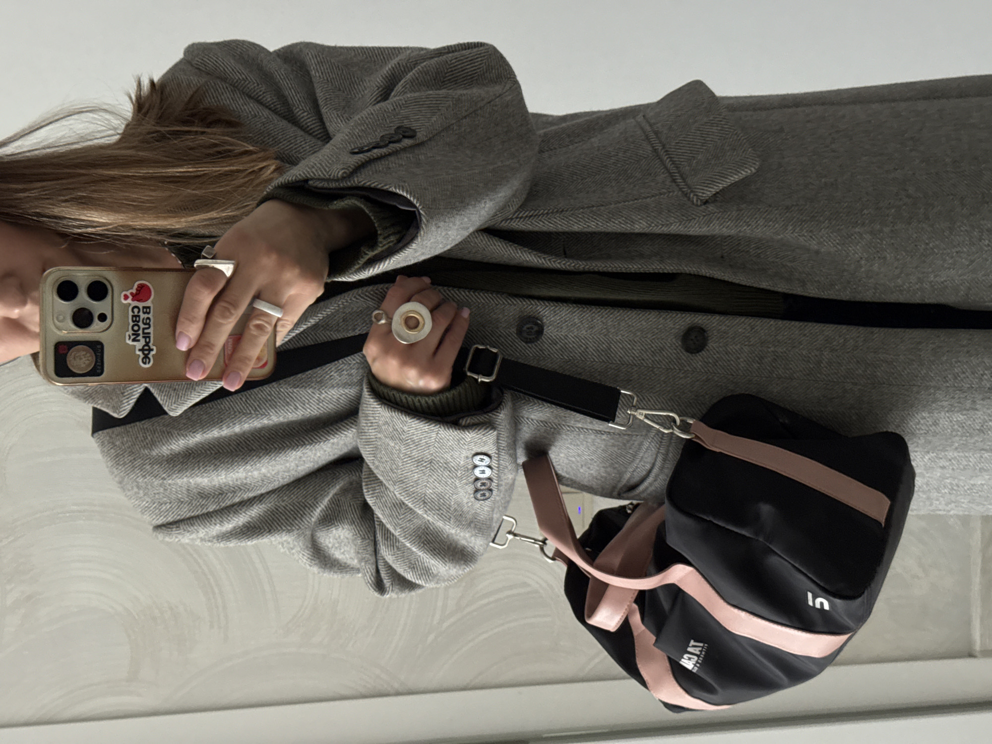 A woman is taking a selfie. She is wearing a grey herringbone coat and an olive green sweater. She has three rings on her fingers, and is holding a smartphone with a sticker on the back in Russian. A black and pink duffel bag hangs from her shoulder. The scene is likely indoors, given the indirect light and reflective floor. There is no conclusive evidence that the location is Sochi, Russia.