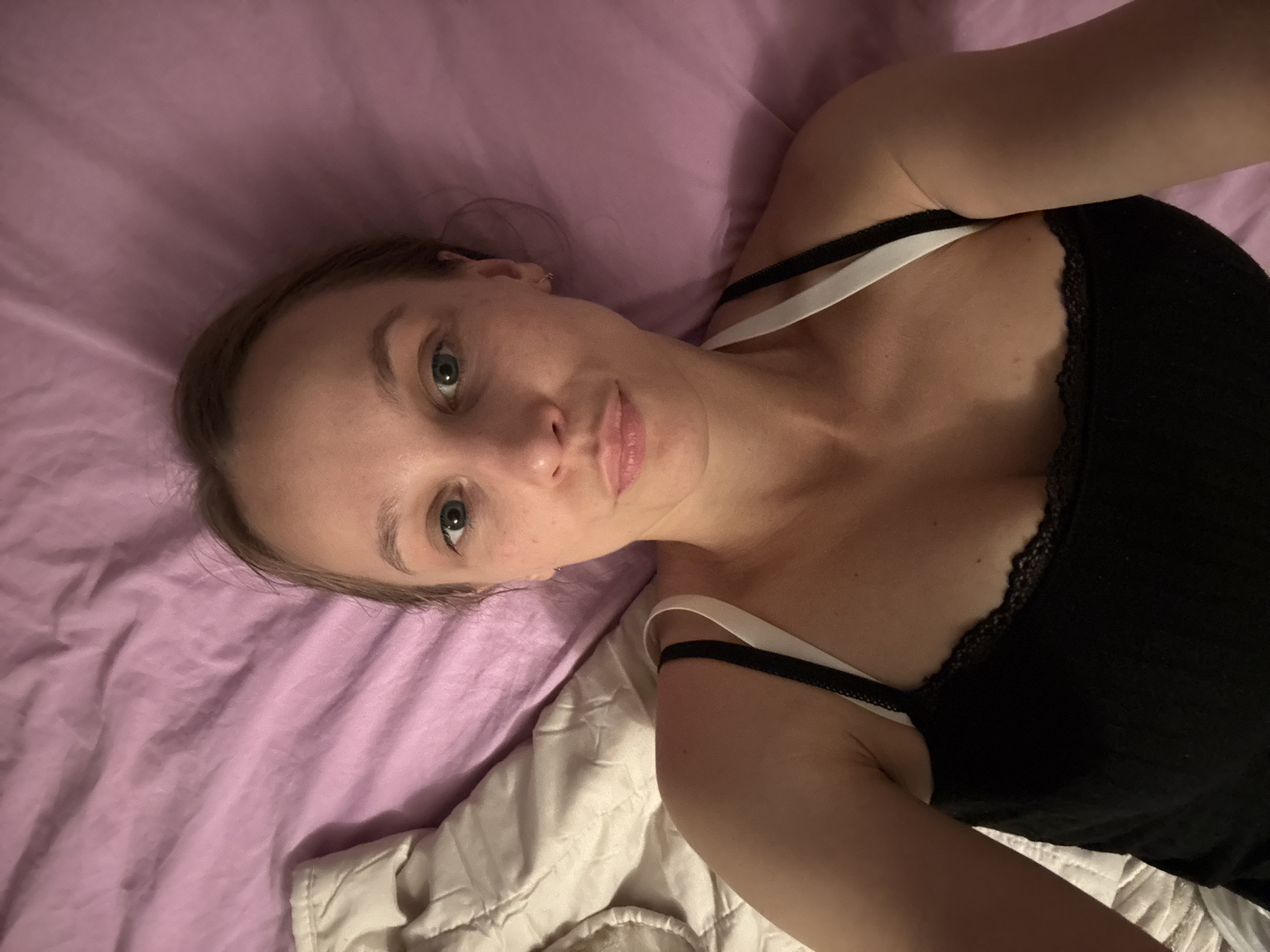 The image shows a close-up, high-angle selfie of a young Caucasian woman lying in bed, with her head tilted slightly to her left. She has fair skin, light brown hair pulled back, and bright green eyes. She is wearing a black tank top with white straps and black lace trim. Her lips are slightly parted, and she appears to be looking directly at the camera. The background consists of a wrinkled pink duvet and a cream-colored textured blanket. The scene appears to be an indoor bedroom setting, possibly in Überherrn, Germany, though this location cannot be confirmed from the image alone.