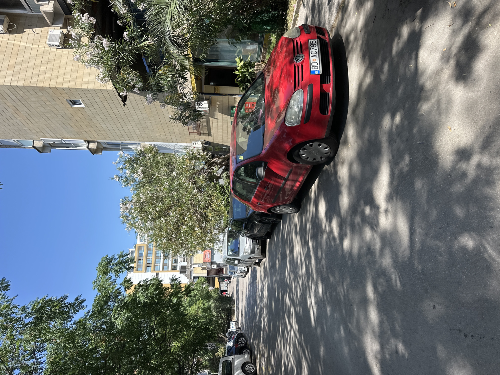The image depicts a street scene in Budva, Montenegro. A red Volkswagen car, bearing a license plate "BD AC795", is parked along the right side of the road. Other parked cars are visible further down the street. Buildings line the street, with trees and vegetation present. The sky is clear and blue, suggesting a sunny day. The scene appears to be a typical residential or commercial area.