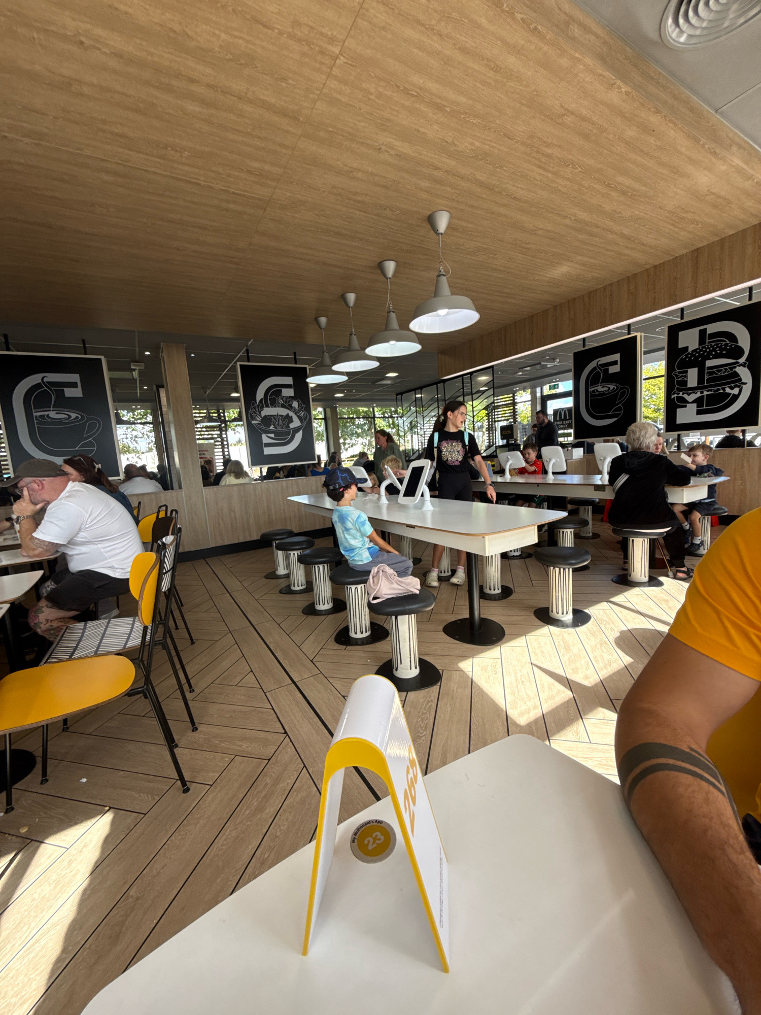 The image depicts the interior of a McDonald's restaurant, likely located in Colchester, United Kingdom, evidenced by the branding and typical McDonald's interior design elements such as the wooden-patterned ceiling, the characteristic table and seating arrangements, and the large wall graphics featuring stylized "C" logos with icons of a coffee cup and a burger.
In the foreground, a white table with a yellow and white sign displaying the number "23" is visible. To the right, a person's tattooed arm, with dark bands on the forearm, is resting on the table. The flooring is composed of wooden planks laid in a herringbone pattern.
Further into the restaurant, several tables and chairs are occupied by patrons. On the left, a man wearing a white t-shirt and a cap is seated at a table. In the middle ground, a young boy in a blue t-shirt and a hat sits at a table with black and white stools. Behind him, a young woman with a backpack is standing and appears to be interacting with a tablet. Several other individuals are seated at tables in the background, some of whom are children. The restaurant is illuminated by overhead lighting, and natural light enters from windows in the background, suggesting it is daytime. The overall atmosphere is that of a busy, casual dining environment.
