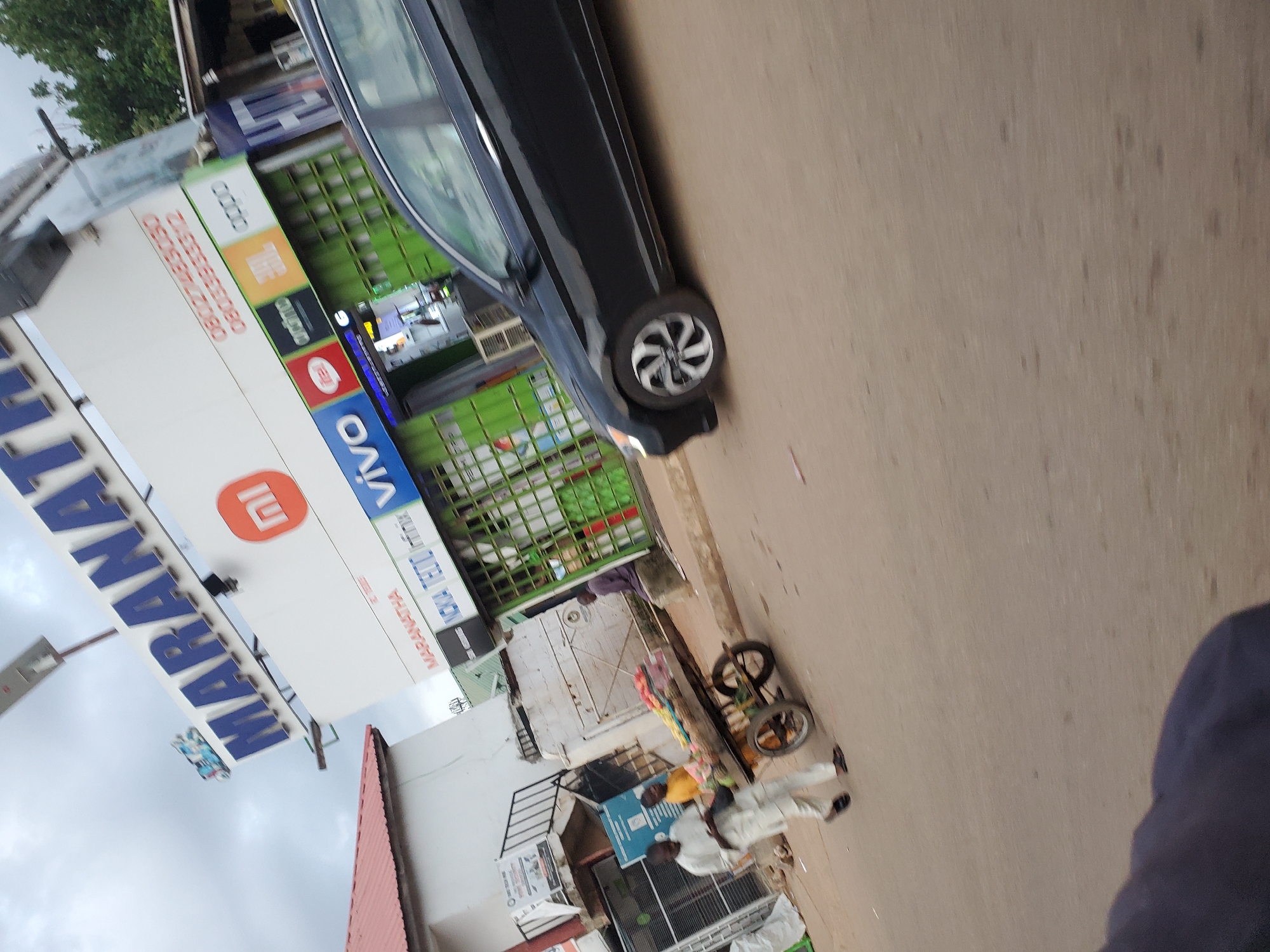 The image captures a street scene in Bukuru, Nigeria, featuring a storefront with prominent signage for "MARANATHA" in large blue lettering against a white background. Below the main sign, various brand logos like "mi," "NOKIA," "TECNO," "Infinix," and "VIVO" are visible, indicating a store selling mobile phones and electronics. The store itself has a green, grate-like facade.
In the foreground, a black car is partially visible, angled towards the right side of the frame. To the left of the car and in front of the store, a man dressed in white is pushing a cart laden with fruits. Another person, wearing a yellow shirt, is standing near the cart. Further down the street, a man is seated. The pavement is visible, and the overall environment appears to be an urban commercial area with buildings and a street. The sky is overcast.