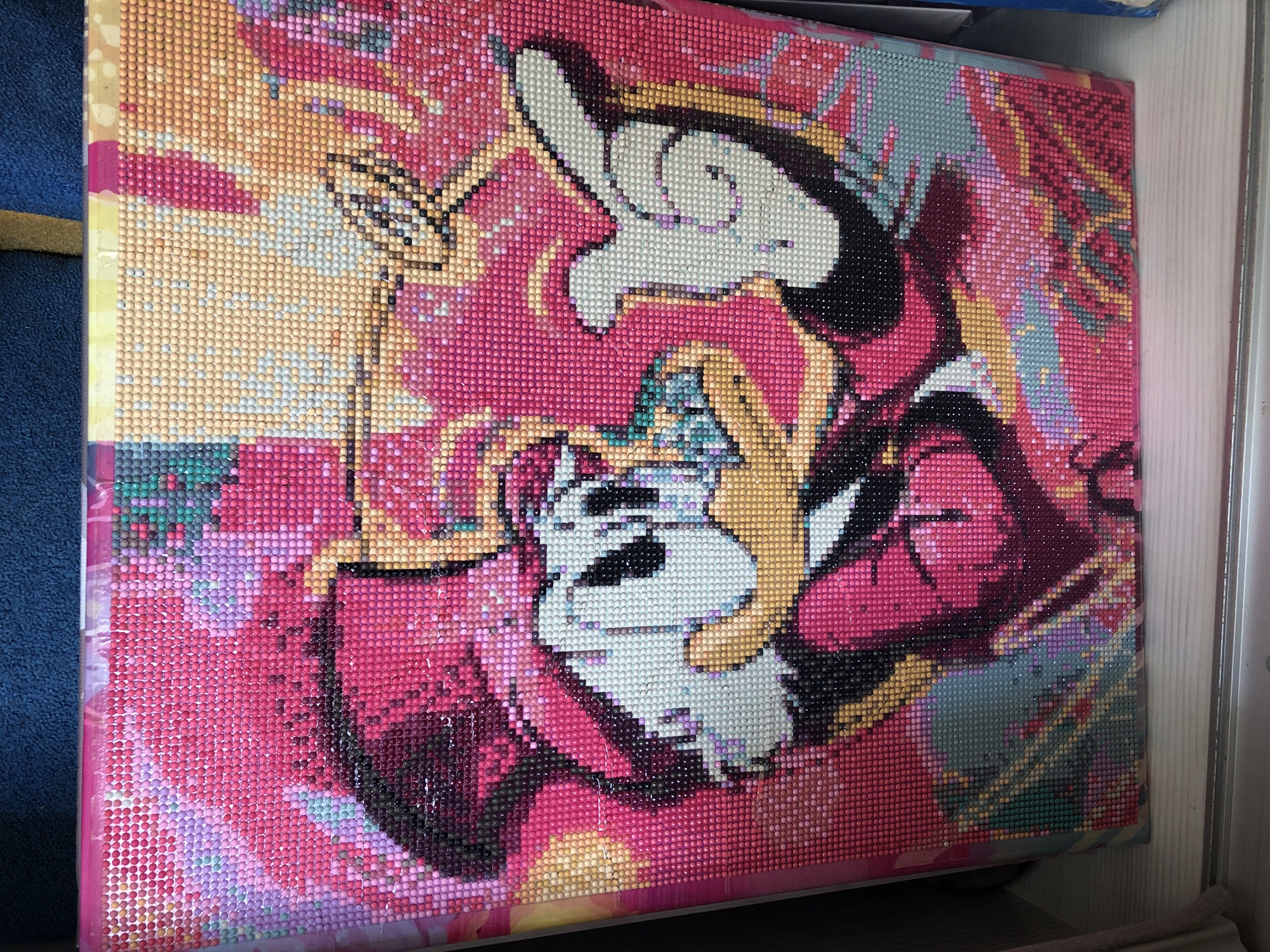 The image depicts a partially completed diamond painting of Mickey Mouse holding a cat, with a primarily pink and yellow color scheme. The painting appears to be on a canvas stretched over a frame. The edges of the frame are visible. The background includes glimpses of a blue surface, a yellowish object, and a white door frame. The image is not in Noviy, Russia as there is no information present to place it there.