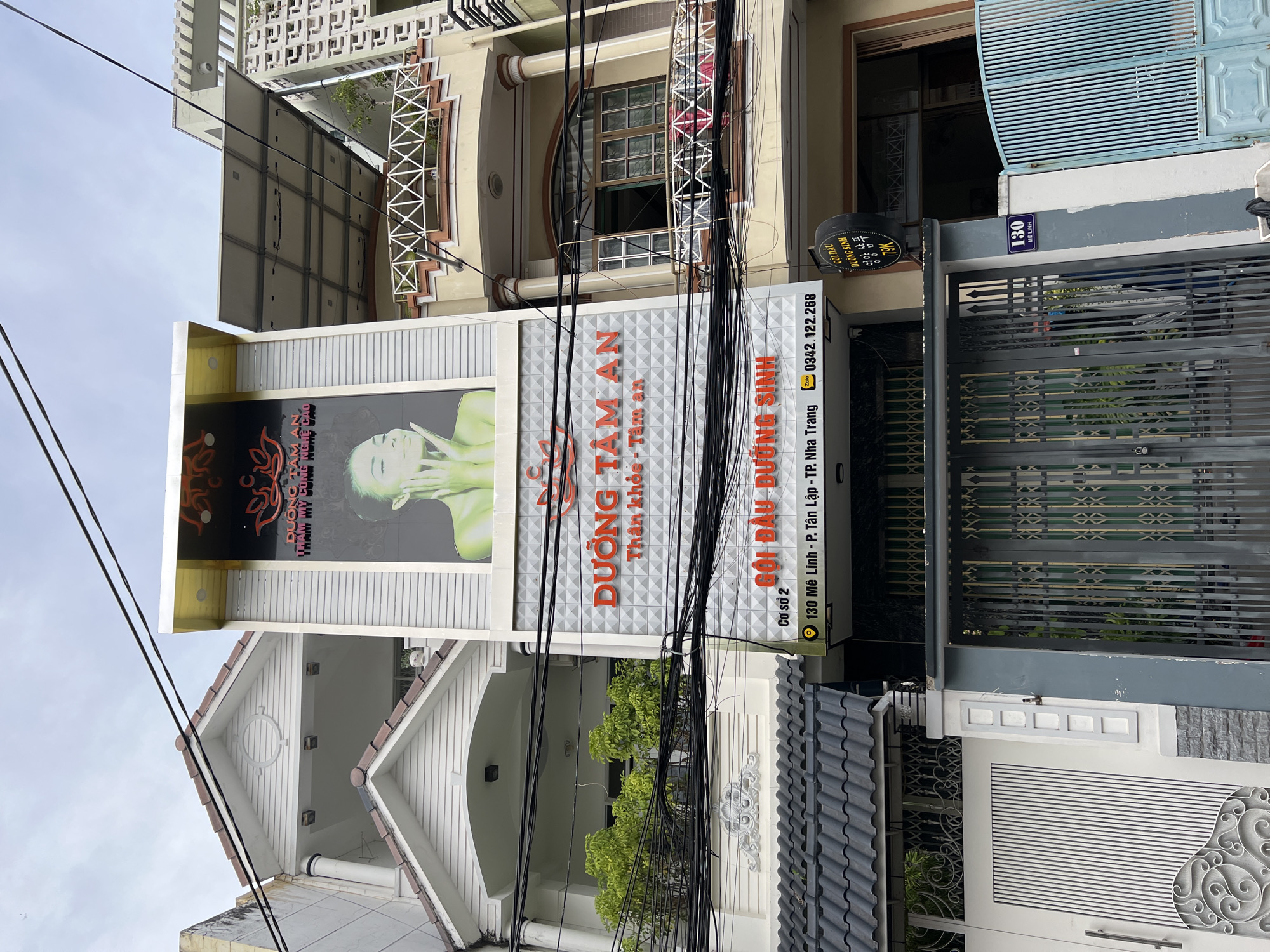 This is a photograph of a multi-story building in Nha Trang, Vietnam, which appears to be a spa or beauty salon. The building features a prominent facade with signage. On the upper part of the building, a large banner displays an image of a woman's face with green skin, along with Vietnamese text. Below this banner, another sign in orange lettering reads "DUONG TAM AN Than khoe - Tam an". Further down, another orange sign says "GOI DAU DUONG SINH", indicating a hair washing and childbirth service. A smaller sign below that provides an address: "Co so 2 130 Me Linh - P. Tan Lap - TP. Nha Trang" and a phone number "0342.122.268". The building is adorned with various architectural details, including balconies with decorative railings, and the facade is decorated with geometric patterns. Electrical wires are visible strung across the front of the building. In front of the building, a metal gate is visible, as well as a sign with the number "130" and "Me Linh". The sky above is overcast.
