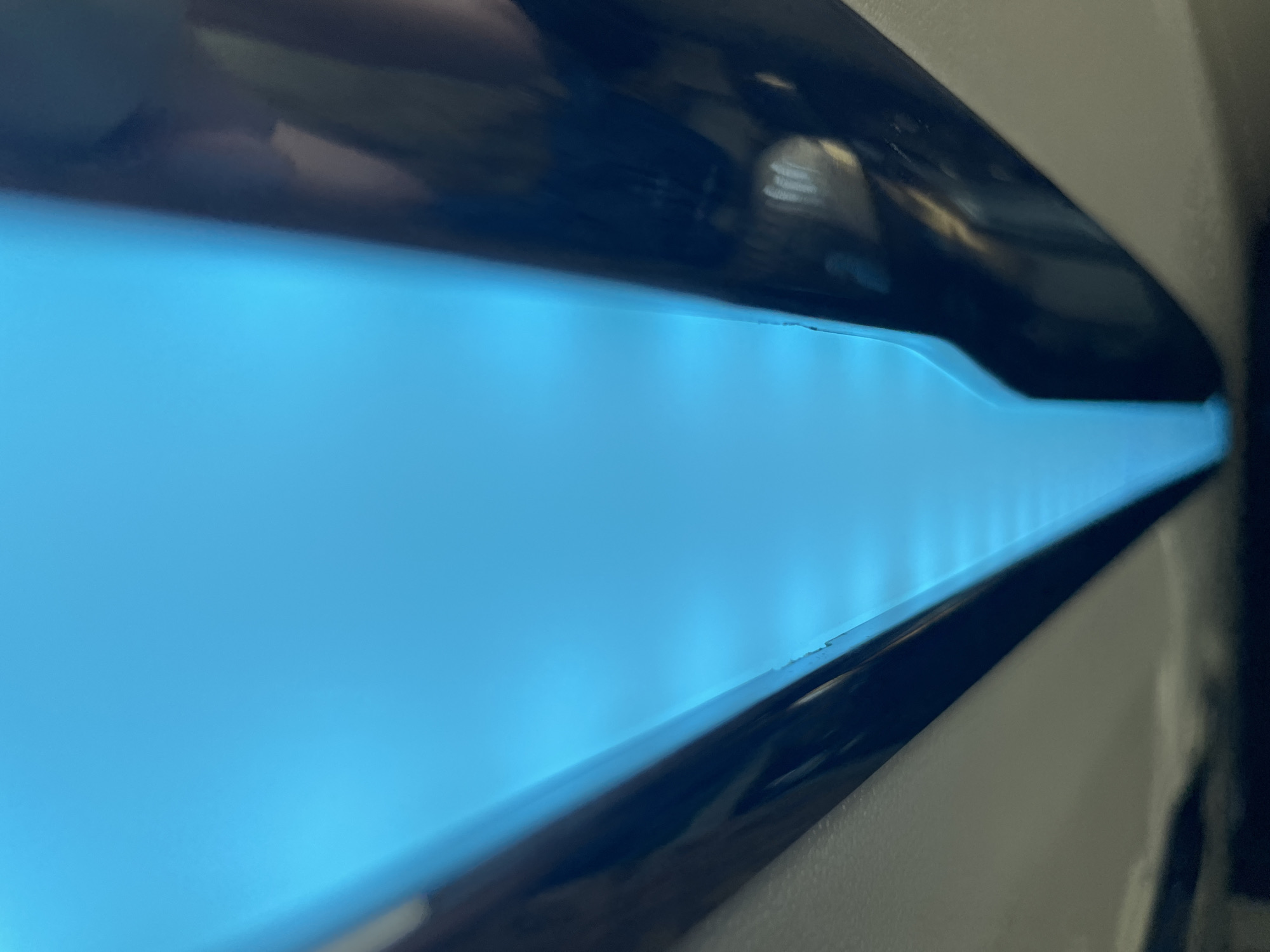 The image shows a close-up, abstract view of a bright cyan light emanating from what appears to be the interior of a device. The light is diffused and forms a V-shape, with dark, reflective surfaces on either side. The surfaces have a glossy finish, and subtle reflections of what could be a human hand and some internal components are visible. The overall impression is of a high-tech or industrial setting, but the specific location within Dong Nai City, Vietnam, cannot be determined from the visual information provided.