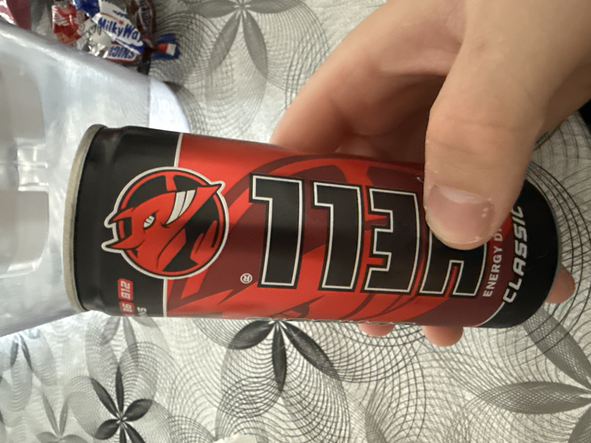 A hand holds a red and black "Hell" energy drink can. The can features a red devil logo within a circle. Part of a clear plastic container and a "Milky Way" candy wrapper are visible in the background. The background also shows a tablecloth with a gray floral pattern. The location is not discernible from the image.