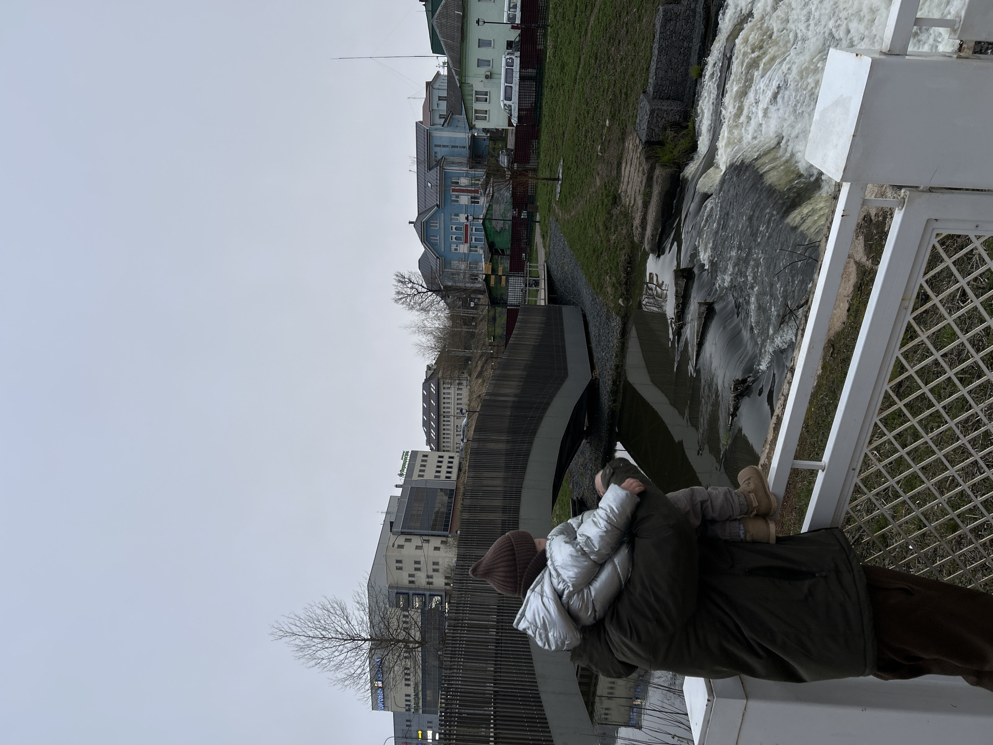 The image, taken in Klin, Russia, depicts an adult holding a child, both observing a river or canal under an overcast sky. The individuals are positioned on the right side of the frame, standing behind a white metal railing with a grid-like pattern. The adult wears a dark olive green winter coat and a brown knitted beanie, while the child is dressed in a shiny silver puffer jacket and light brown boots. They appear to be looking towards the left, across the waterway.
The river features a section of rushing, white water on the right, indicating rapids or a small waterfall. This water then flows left, becoming calmer as it passes under a large, curved concrete structure, likely a bridge or part of a dam. The riverbanks are reinforced with gabion walls and patches of green grass.
Along the far bank of the river, a mix of architectural styles is visible. Closer to the foreground on the left are several colorful, older buildings, including a distinctive blue house and a lighter green one. A white bus or van with the letter "N" on its side is parked nearby. Further along the river, more modern, multi-story buildings with reflective windows are present. On one such building, a blue sign with "FORUM" is visible, and on another, a green sign partially shows what appears to be "СТРОЙКА" (construction) and possibly "МЕБЕЛЬ" (furniture). Bare trees scattered throughout the scene suggest either late autumn or early spring. The muted lighting from the cloudy sky contributes to a somewhat cool and subdued atmosphere.