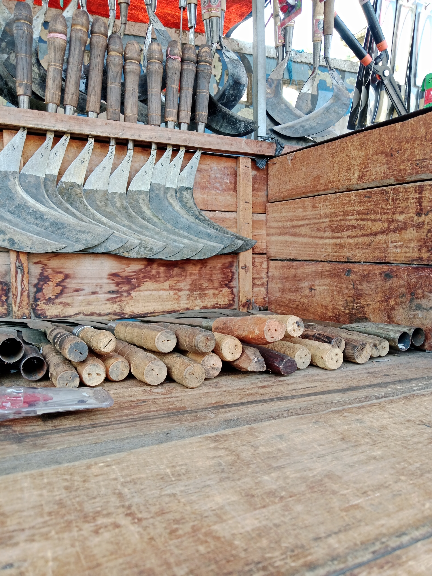 The image shows a close-up, low-angle shot of a collection of farming tools, likely scythes and machetes, displayed on wooden surfaces. The tools are arranged in two distinct groups. The upper group consists of several scythes hanging from a wooden rack, their curved blades pointing downwards. The lower group comprises numerous wooden handles of various tools lying horizontally on a wooden plank. The background is dominated by weathered wooden planks, suggesting an outdoor market stall or a storage area. The lighting appears natural, highlighting the textures of the wood and metal. No people are visible in the frame. The scene is likely situated in a traditional market or workshop in Kediri City, Indonesia, where such tools are commonly sold or manufactured for agricultural use.