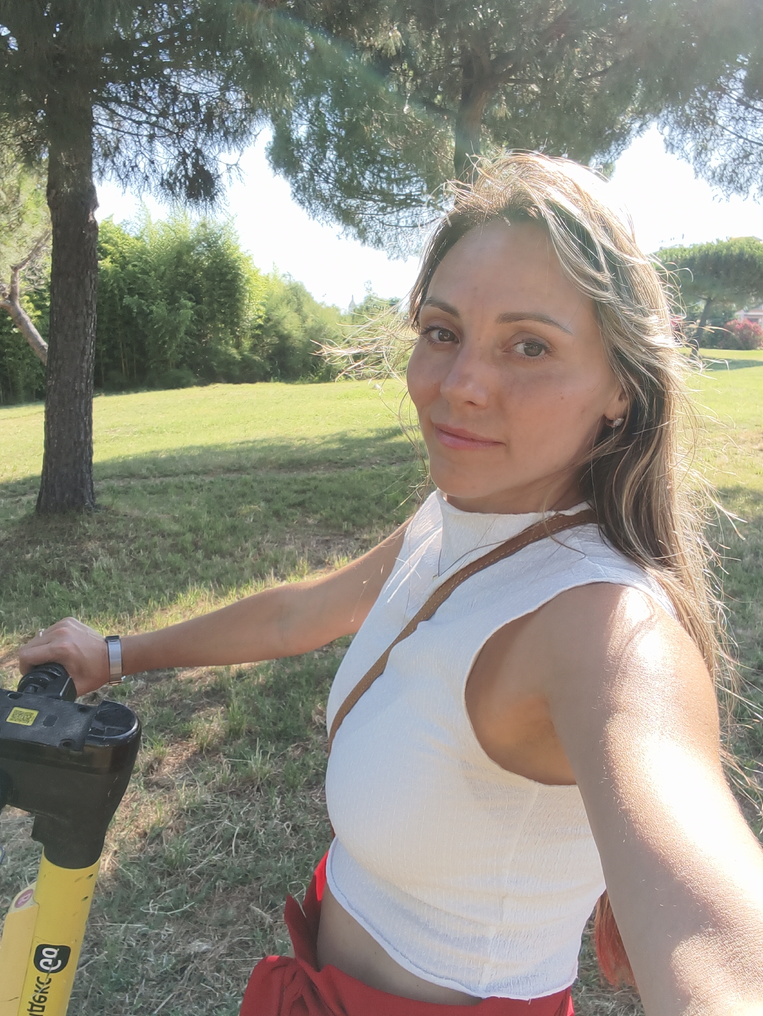 A woman with blonde hair and light skin is taking a selfie outdoors. She is wearing a white, sleeveless, crop top and red shorts. She is holding the handlebars of a yellow and black electric scooter. The background is a park-like setting with green grass, trees, and bushes. The sun is shining, casting shadows on the ground. The location is not specified as Sirius, Russia.