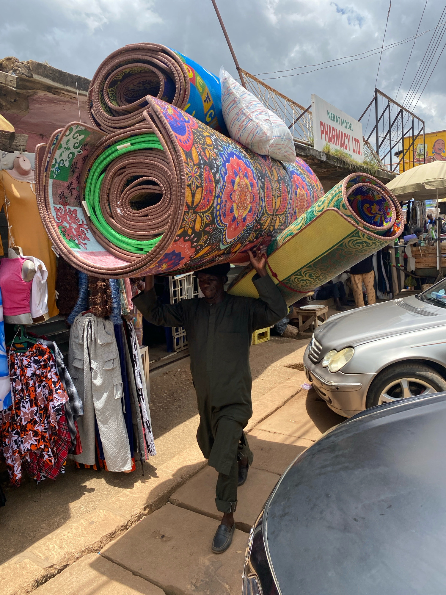 A man in a dark green uniform walks along a dusty street carrying several rolled-up mats on his head. The mats are of various colors and patterns, including a blue mat with letters and a colorful floral-patterned mat. The man is walking past a market stall on the left, which displays clothing on mannequins and hanging racks. Beyond the stall, there are more market stalls and a sign that reads "NERAT MODEL PHARMACY LTD." On the right side of the street, two cars are parked, a silver Mercedes-Benz and a dark gray car. The sky is overcast with clouds, and the ground is made of cracked concrete slabs and dirt. The scene appears to be in Bukuru, Nigeria.