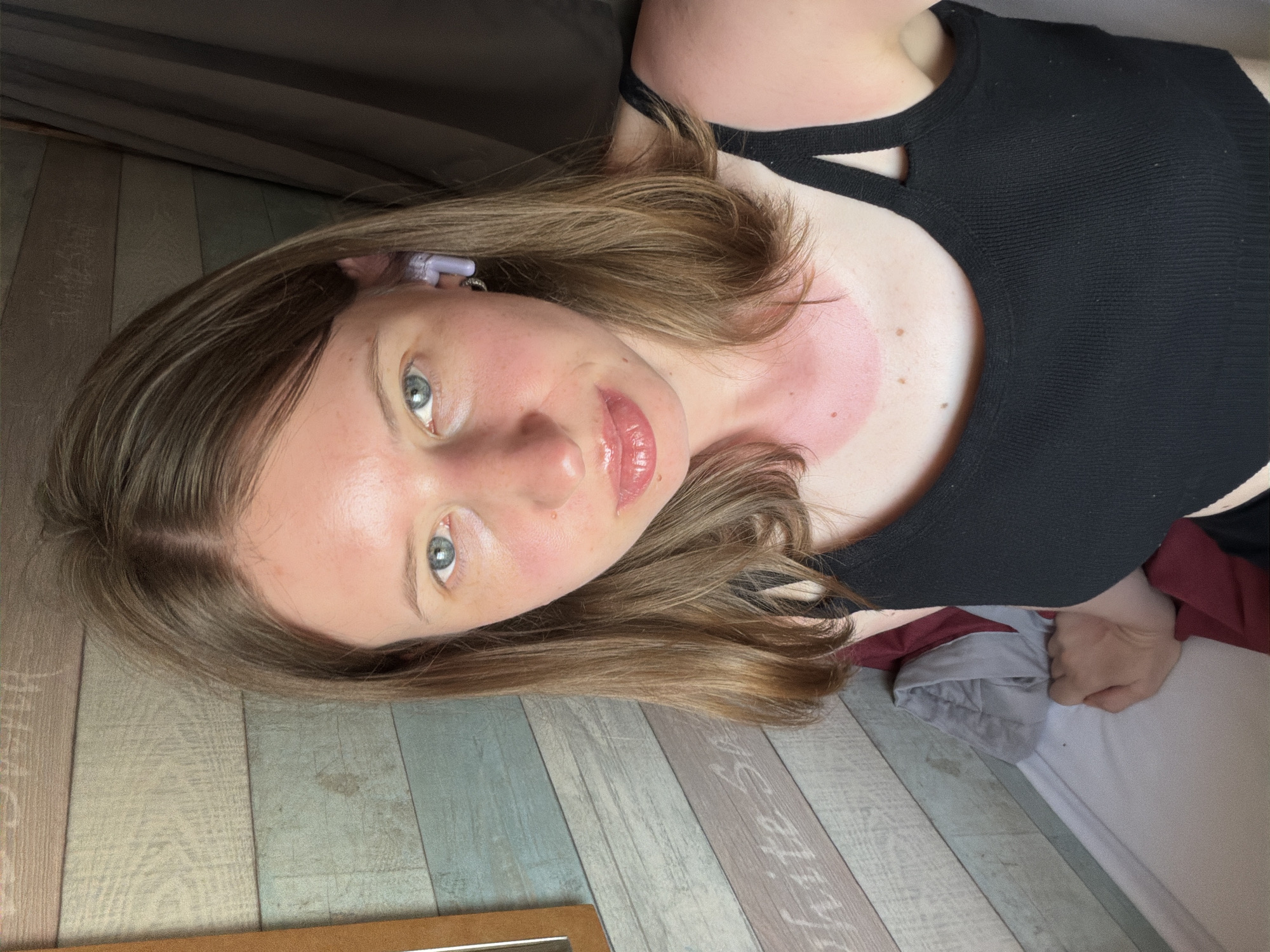 The image is a close-up selfie of a young woman with light brown hair and blue eyes. She is wearing a black tank top and a purple earbud in her right ear. Her skin appears slightly reddened, suggesting a sunburn on her face and neck. She is indoors, with a wall decorated with wooden planks in the background. The lighting is natural, coming from the left. She is looking directly at the camera. There are no other people or significant objects visible in the frame. The location is not specified in the image or provided context.