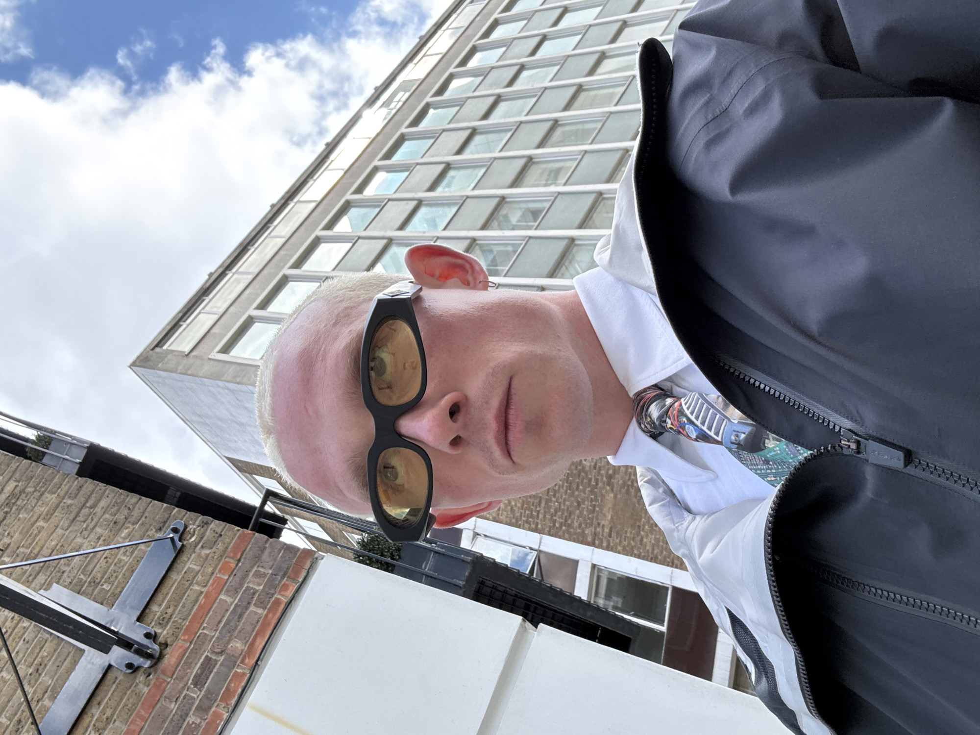 A low-angle, tilted selfie shows a light-skinned person with short, bleached blonde hair, wearing black sunglasses with yellow-tinted lenses. They are dressed in a white button-down shirt with a multi-colored tie, and a black jacket is partially visible.
Behind the person is a multi-story building with a grid of windows under a cloudy blue sky. The lower part of the scene includes a brick wall with metal fixtures and a white ledge.
The building appears to be located in London, UK, based on architectural style common in the city.