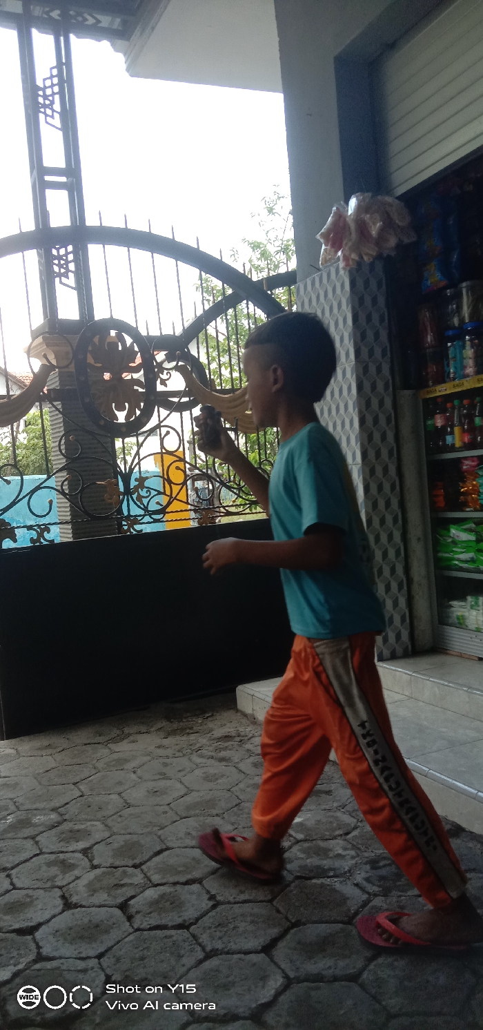 The image depicts a young male walking from right to left on a paved surface in Sebaung, Indonesia. He wears a light blue t-shirt, orange track pants with a grey stripe along the outer leg, and red flip-flops. His short, dark hair is styled with faded sides. He holds a small, dark, irregularly shaped object to his slightly open mouth with his right hand.
To the left, a tall, black wrought iron gate, featuring intricate gold-accented scrollwork and a solid black lower panel, stands partially open. Beyond the gate, green foliage and a distant blue structure are visible. A white building overhang is seen above the gate against a bright, overcast sky.
To the right, the entrance of a commercial establishment, likely a small shop, is visible. It features a patterned grey and white tiled column. Inside the open storefront, shelves stocked with various packaged goods and bottles are visible. Several plastic bags containing items hang above the tiled column. Two white steps lead up to the shop entrance. The ground consists of hexagonal concrete pavers, appearing somewhat damp.
