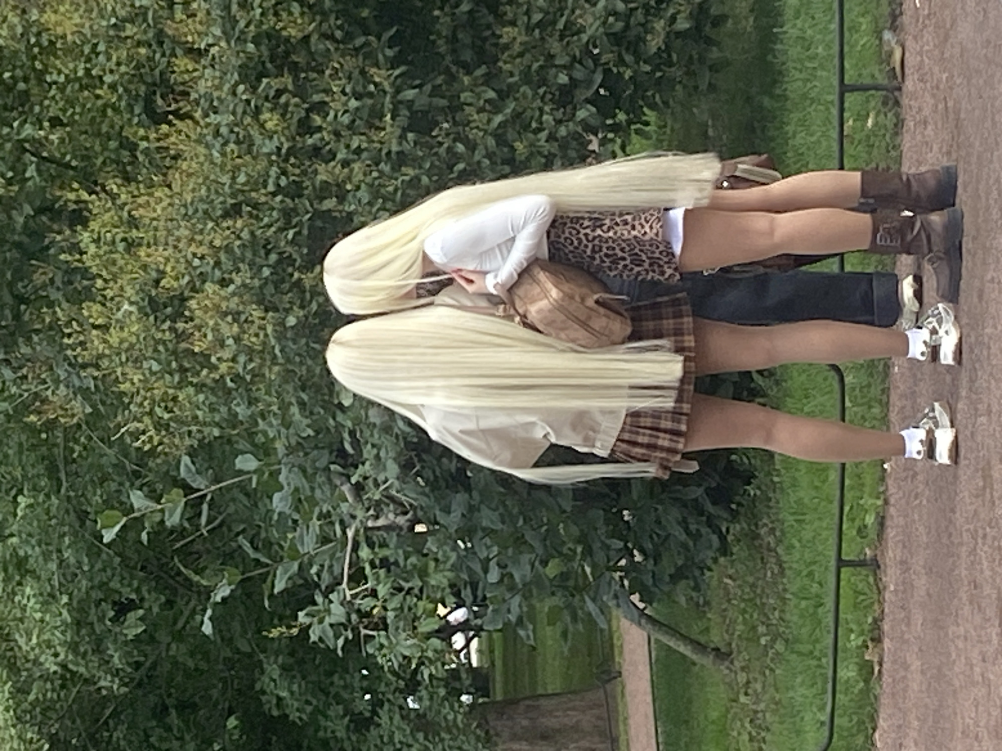 Two individuals with long blonde wigs stand close together, facing away from the camera, in a park setting. The person on the left wears a plaid skirt and a light-colored jacket, while the person on the right wears a leopard print skirt and a white long-sleeved shirt. They are both wearing sneakers. A brown bag is slung over the right person's shoulder. The background consists of lush green trees and foliage, with a path and a grassy area visible. The overall scene suggests a casual outdoor gathering or photoshoot in a park within Saint Petersburg, Russia.