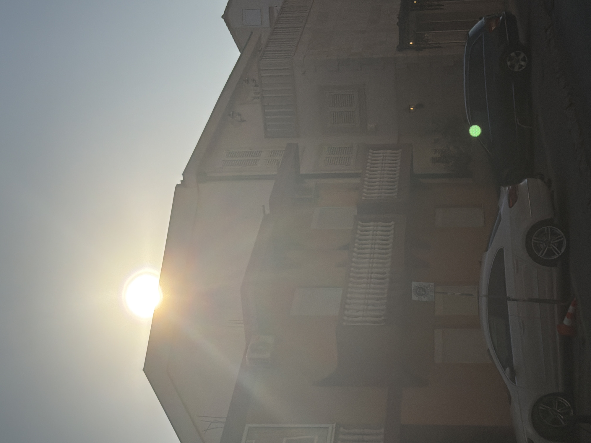 The image depicts a street scene in Budva, Montenegro, under a bright, hazy sun. A multi-story building with cream-colored walls and white balconies dominates the upper portion of the frame. The sun, a bright orb with visible rays, is partially obscured by the haze and positioned above the building's roofline, casting a strong glare. The lower part of the image shows two parked cars on the street. The car on the left is a light-colored sedan, while the car on the right is a dark-colored station wagon. A traffic cone is visible on the ground near the front of the light-colored sedan. The overall impression is that of a sun-drenched, quiet street in a European coastal town, possibly during the late afternoon or early morning due to the low angle of the sun and the hazy light.