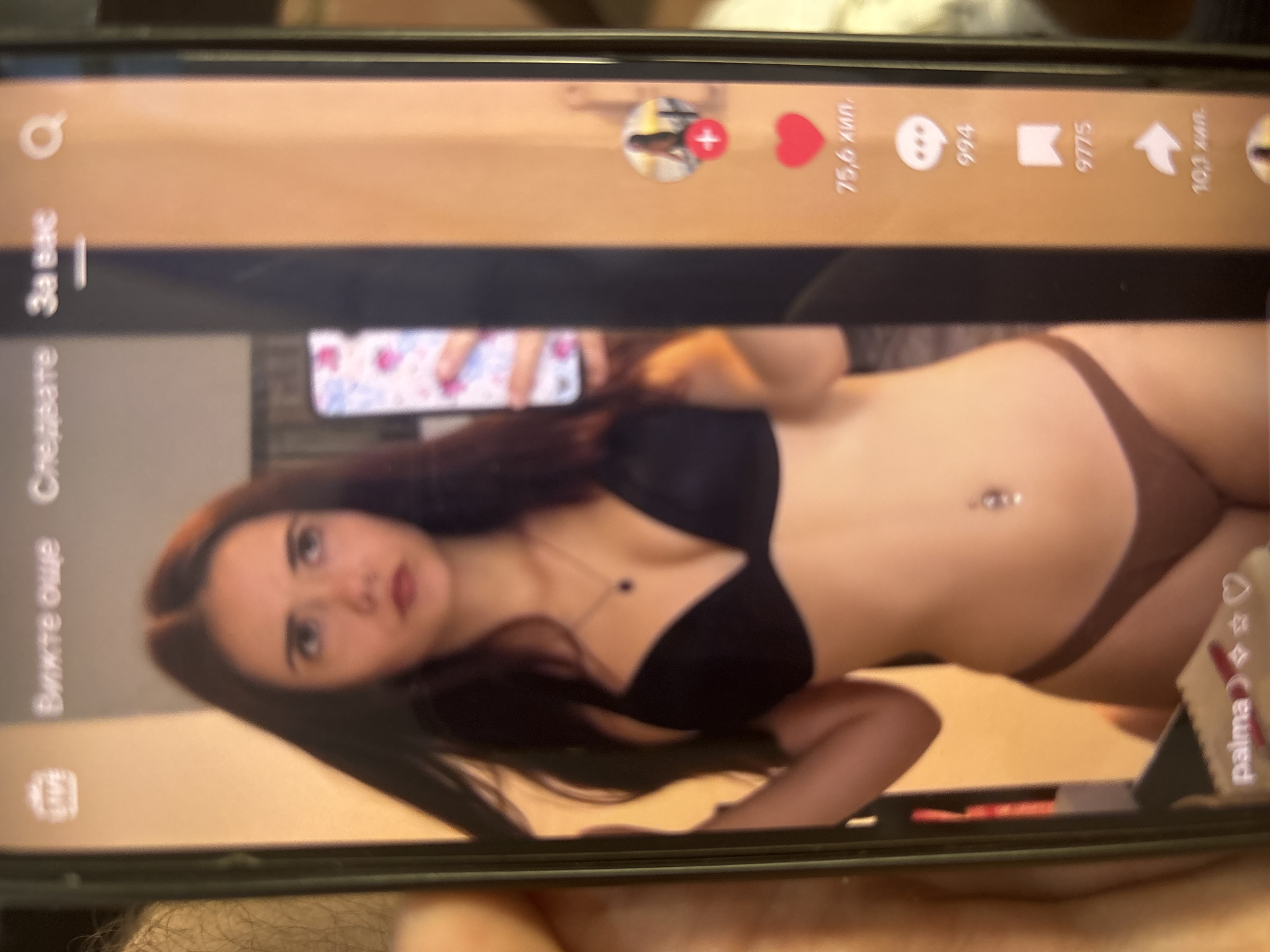 A dark-colored smartphone screen displays a social media interface, featuring a full-body mirror selfie of a young woman. The woman has long, dark hair, a belly button piercing with a silver circular jewel, and wears a black bra and brown/tan underwear. She is holding another smartphone with a colorful, heart-patterned case in her right hand, visible in the mirror's reflection. A silver necklace with a dark pendant is visible around her neck. Her gaze is directed slightly upwards. The background reflected in the mirror appears to be an indoor room with warm, possibly orange or brown-toned walls.
The social media interface overlaid on the image includes several elements:
At the top, partially blurred, are Cyrillic text elements: "LIVE" (likely in English, obscured), "Вижте още" (See more), "Следвайте" (Follow, partially visible), and "За вас" (For you), which is underlined indicating the active tab, followed by a search icon.
Along the right side of the screen, various engagement metrics are displayed: a circular profile icon (likely of the woman in the image) with a red plus sign, a red heart icon followed by "75,6 хил." (75.6 thousand) likes, a speech bubble icon followed by "994" comments, a bookmark icon followed by "9775" saves, and a share arrow icon followed by "10,3 хил." (10.3 thousand) shares.
At the bottom left, partially obscured, is the username "palma" followed by emojis including a moon, star, and heart.
The smartphone is being held in a hand with visible hair on the arm, suggesting it belongs to a male individual. The background behind the phone is out of focus. The on-screen text, written in Bulgarian Cyrillic, indicates the image's origin is Blagoevgrad, Bulgaria.