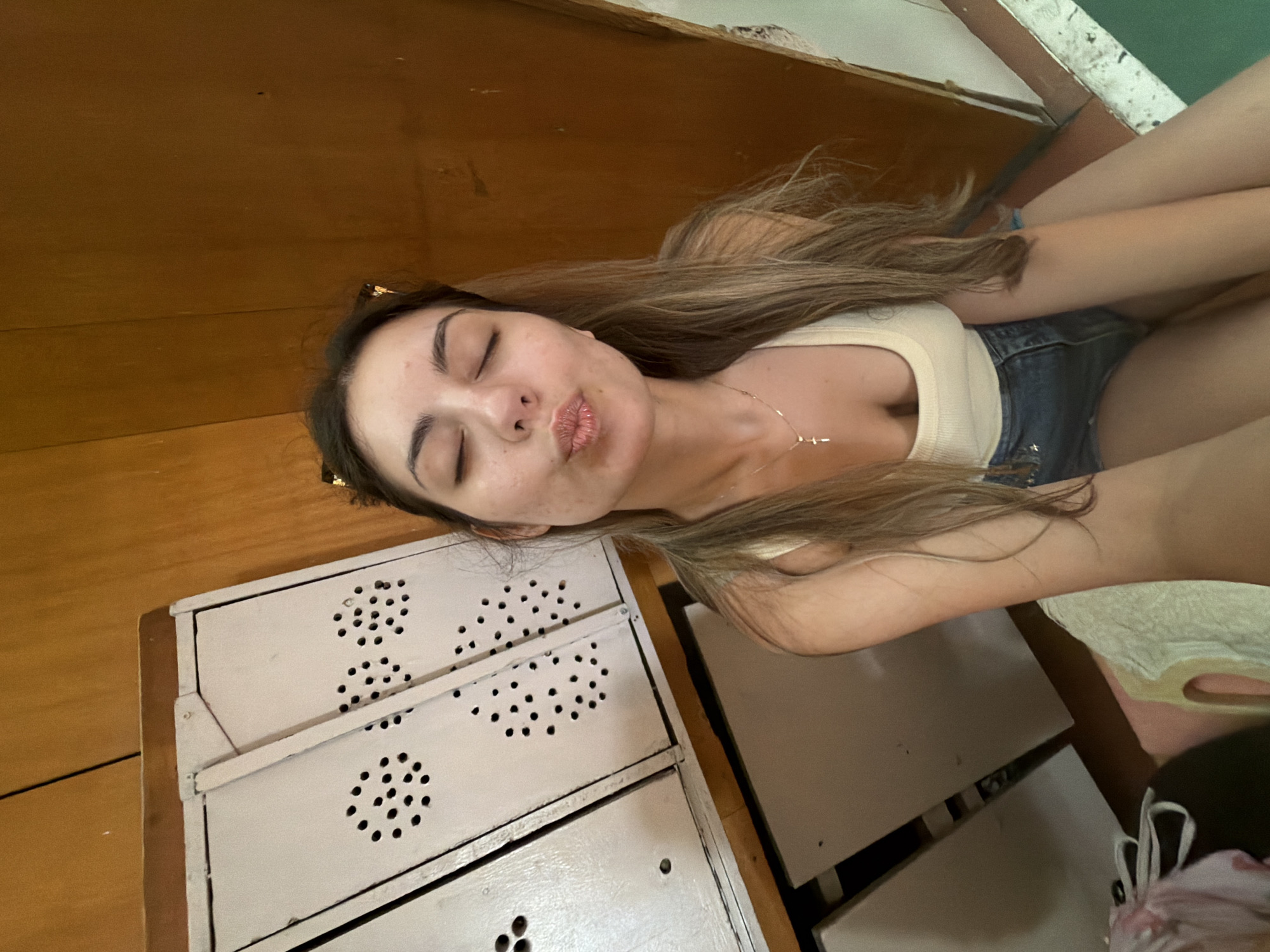 A young woman with long brown hair, wearing a beige tank top and denim shorts, is captured in a selfie with her eyes closed and lips pursed in a kissing gesture. She is wearing a delicate gold necklace with a cross pendant. The background reveals a wooden wall and a white cabinet with circular ventilation holes. The scene appears to be indoors, possibly in a locker room or an old storage area, and is devoid of any specific identifiable landmarks of Burgas, Bulgaria.