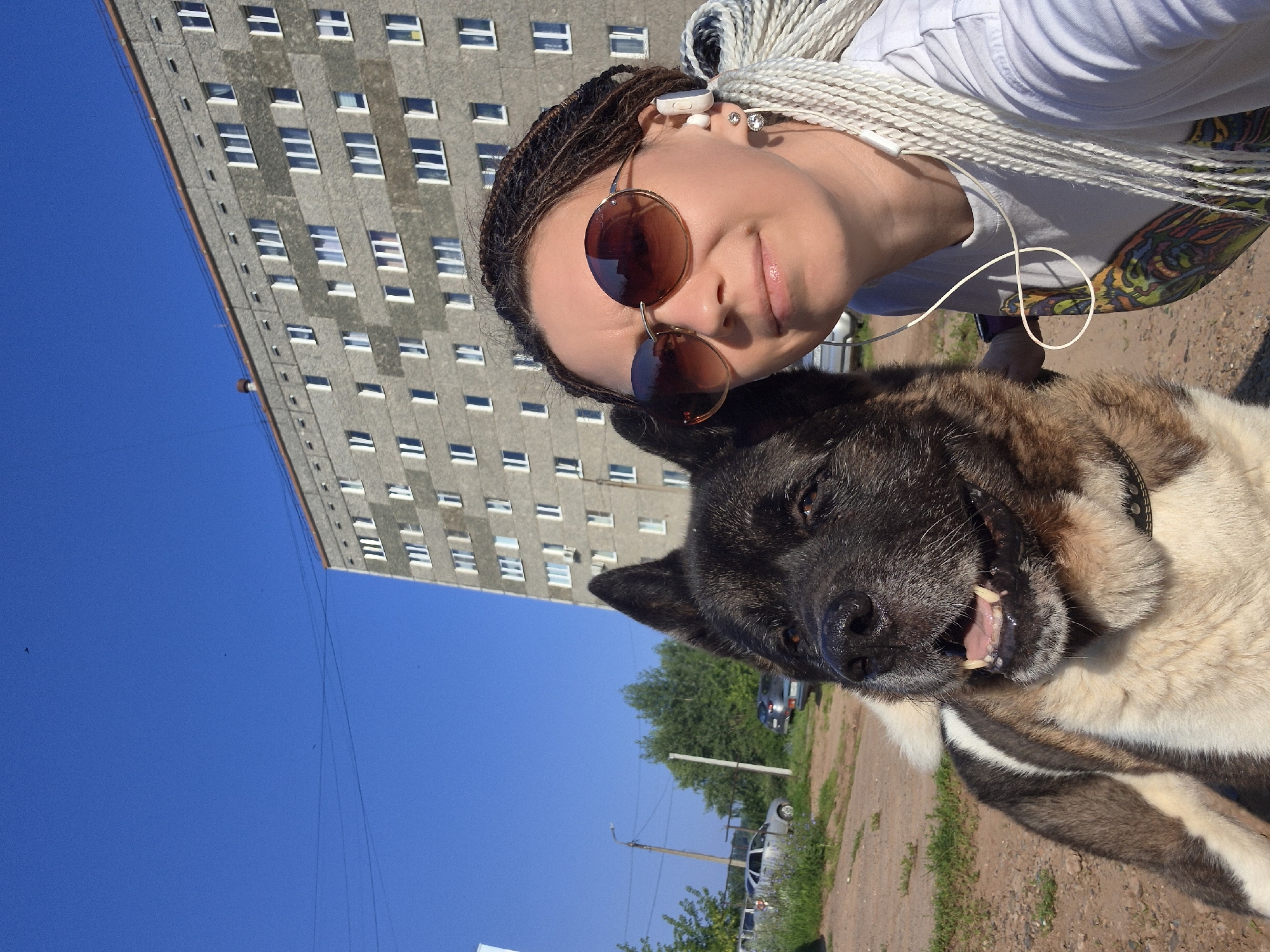 In an outdoor urban setting, a woman with long, braided white hair and round, brown-tinted sunglasses poses for a selfie with a large, dark-haired dog. The woman wears a white t-shirt with a colorful graphic print and white wireless earbuds. She is positioned to the right of the frame, with the dog's head and chest filling the left side. The dog, an American Akita breed, has dark fur on its head and back, and lighter fur on its chest and legs, with prominent ears and a black collar.
Behind them, a tall, gray, multi-story apartment building with numerous windows dominates the background. The sky above is a clear, vibrant blue. To the left of the building, trees and a streetlamp are visible, along with parked cars. The ground in the foreground appears to be unpaved, with dirt and patches of grass. The overall impression is a sunny day in a residential area.